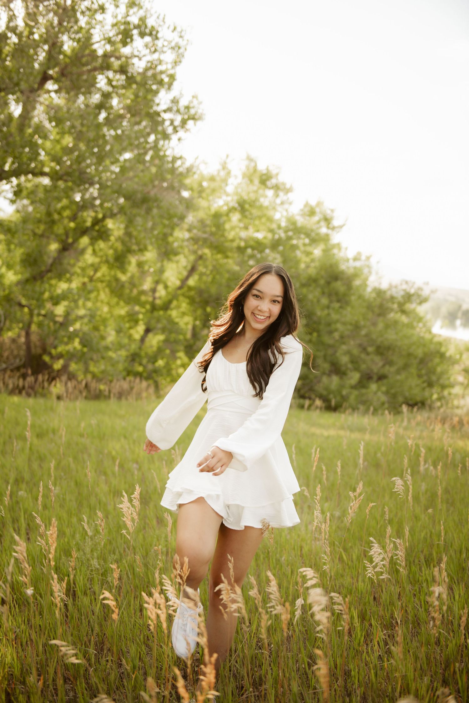 A woman in a flowy white dress twirls joyfully in a sunlit meadow with tall grass and trees in the background.