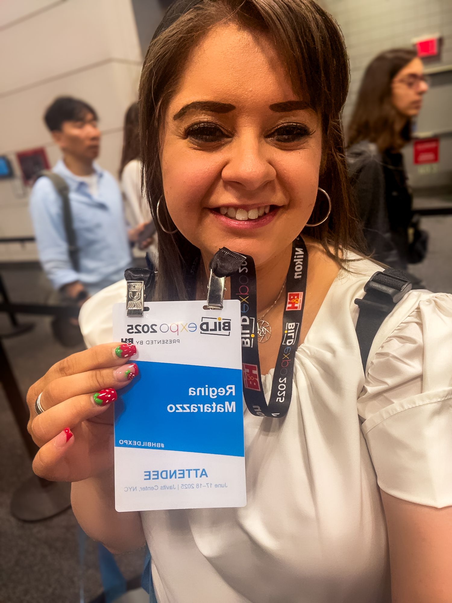 Person smiles while holding up a conference badge with lanyard in an indoor event setting.