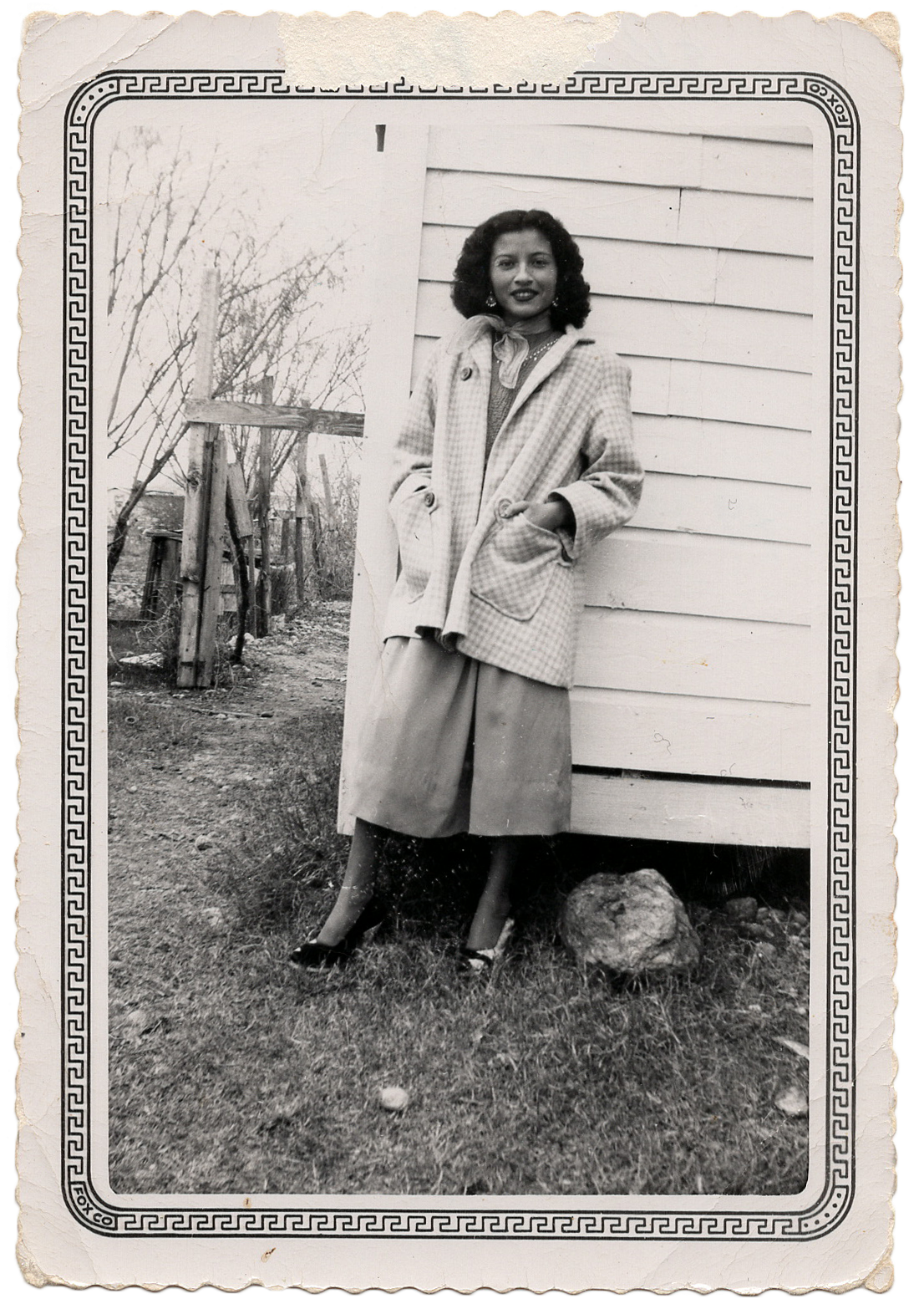 A vintage black and white photograph shows someone wearing a light-colored coat standing outside a house in winter.