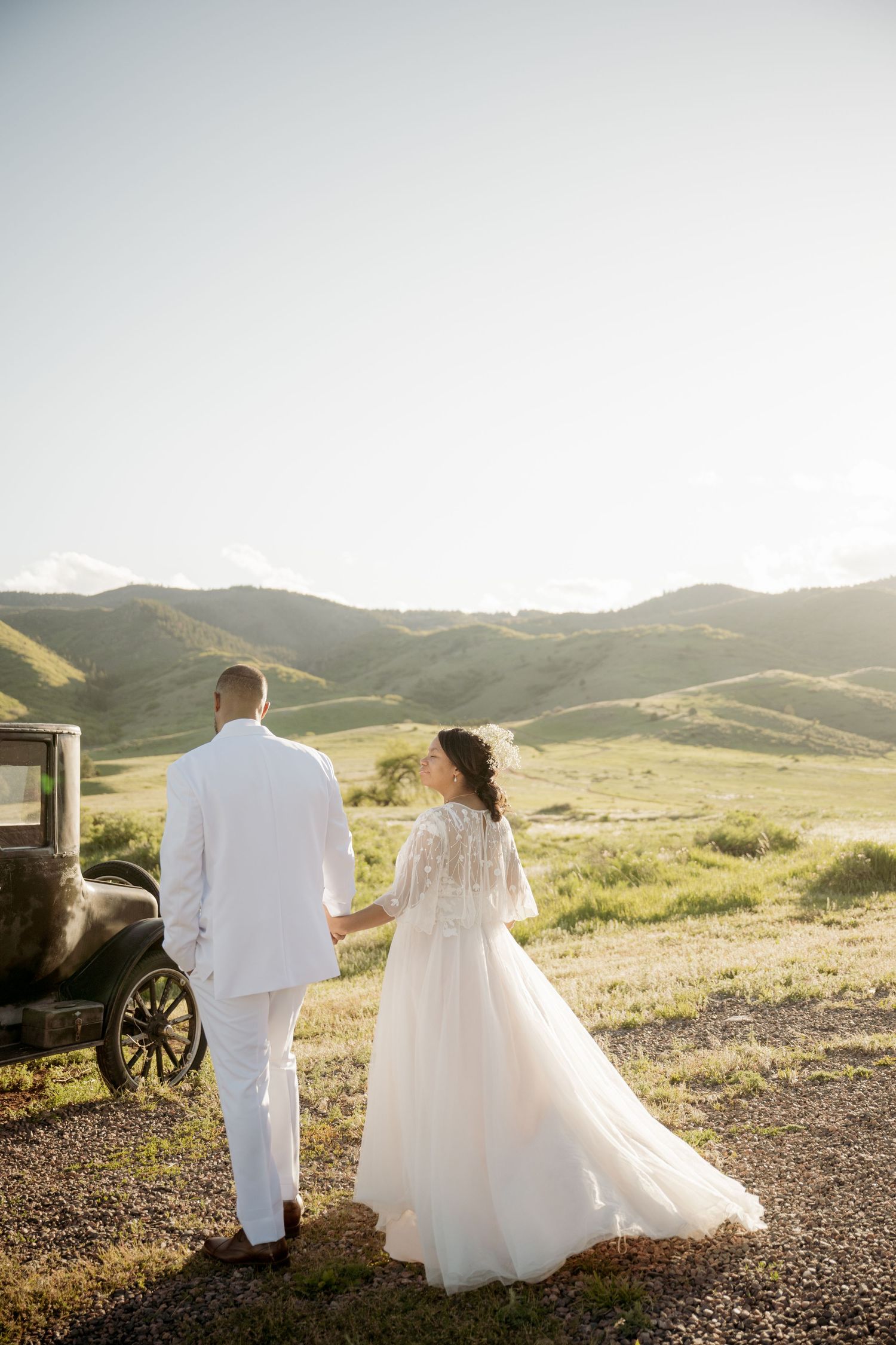 Couple walks hand in hand through golden rolling hills at sunset while wearing wedding attire.
