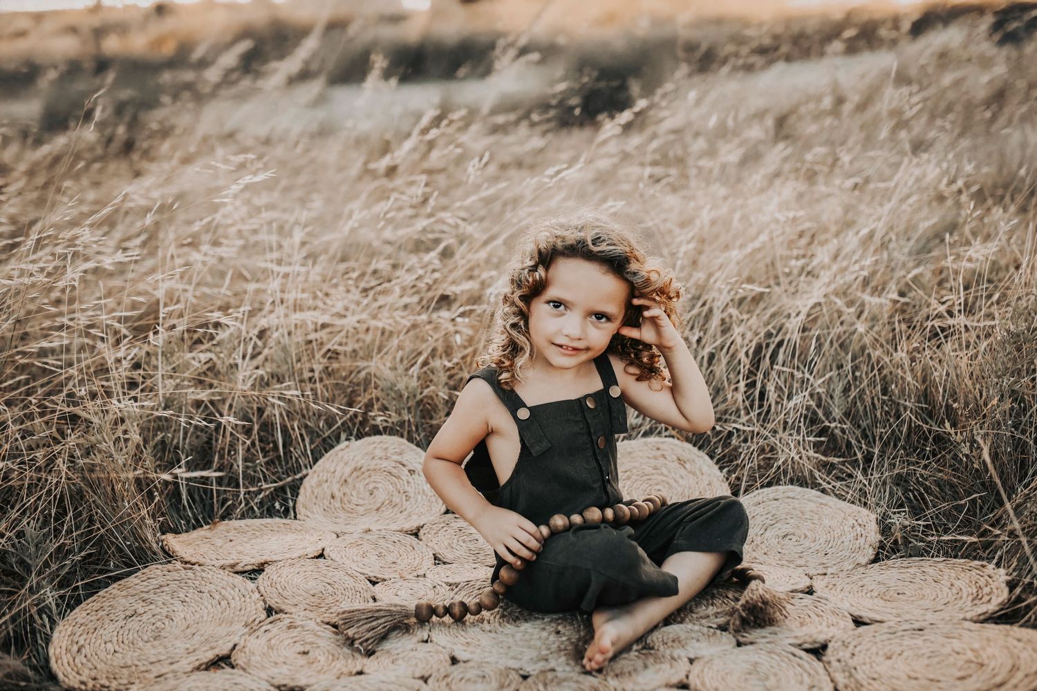 a little girl in Corduroy olive green overalls brushes her curls off of her face as she plays in a field of golden grass at sunset