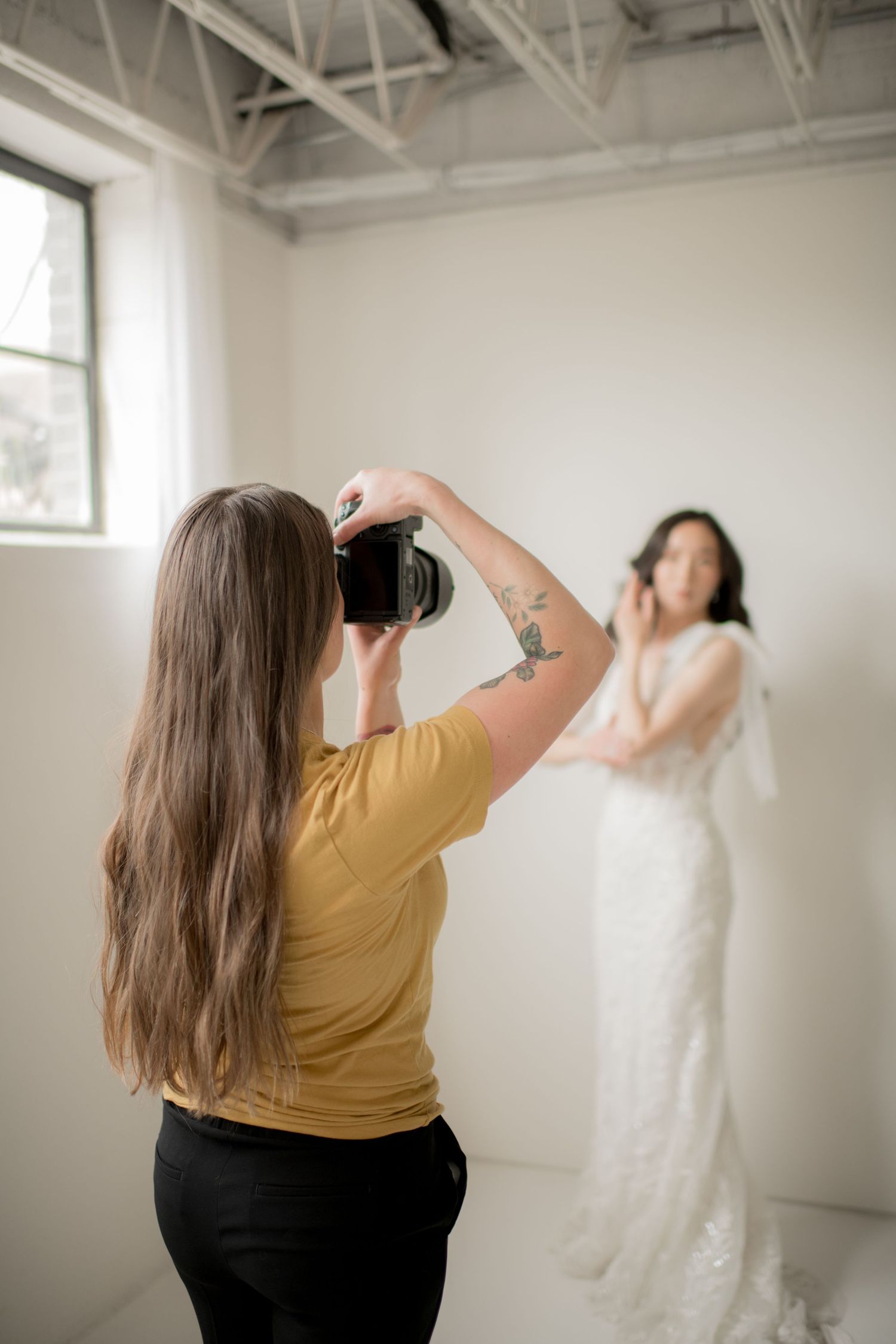 A photographer takes pictures of a white wedding dress hanging in a bright white studio space.