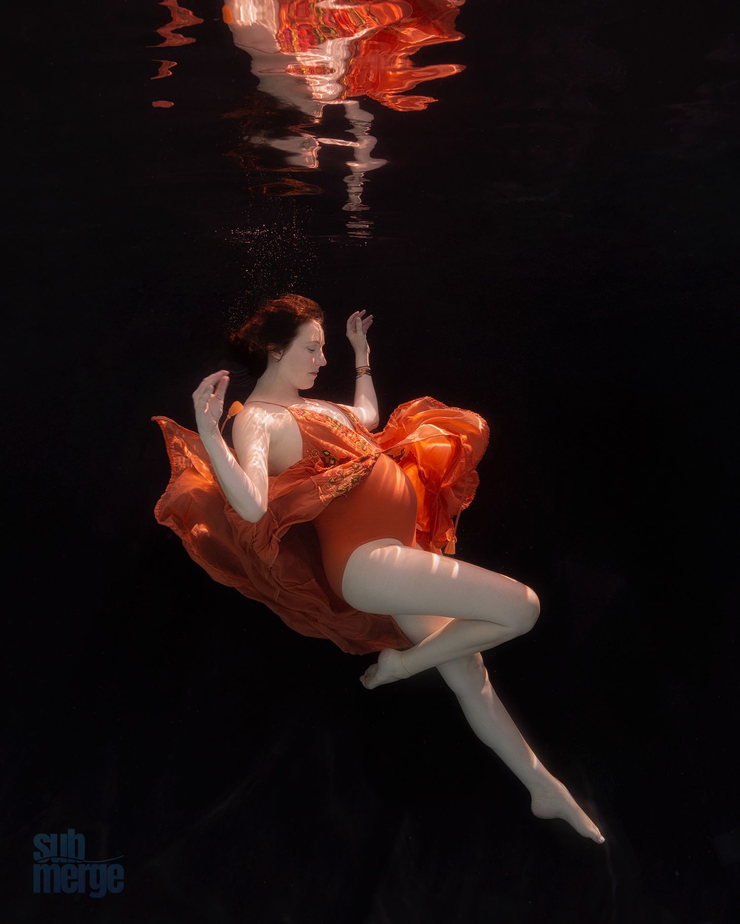 Ethereal underwater portrait of a figure gracefully floating in an orange flowing dress against a dark background.