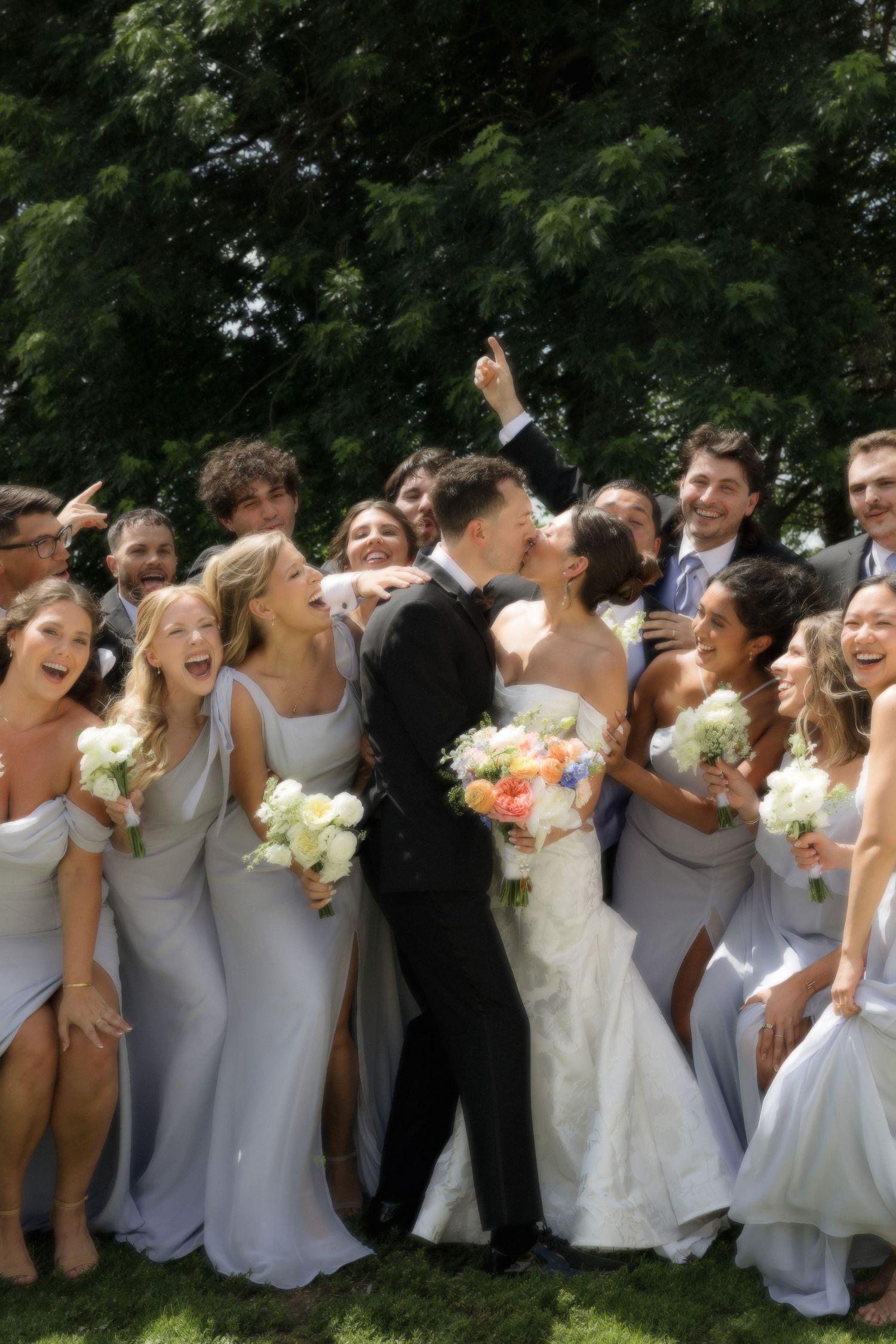 Wedding party in gray bridesmaid dresses and tuxedo gather outdoors for a joyful group photo with flowers.