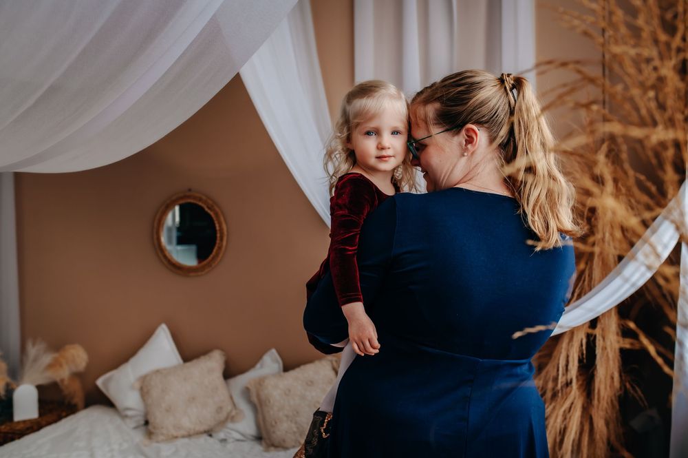 A warm autumn indoor scene with a parent in navy blue holding a small child in burgundy against soft white curtains and decor.
