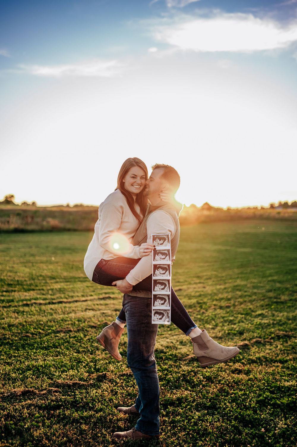 A couple embraces in a sunlit field while holding ultrasound photos during a romantic pregnancy announcement photoshoot.