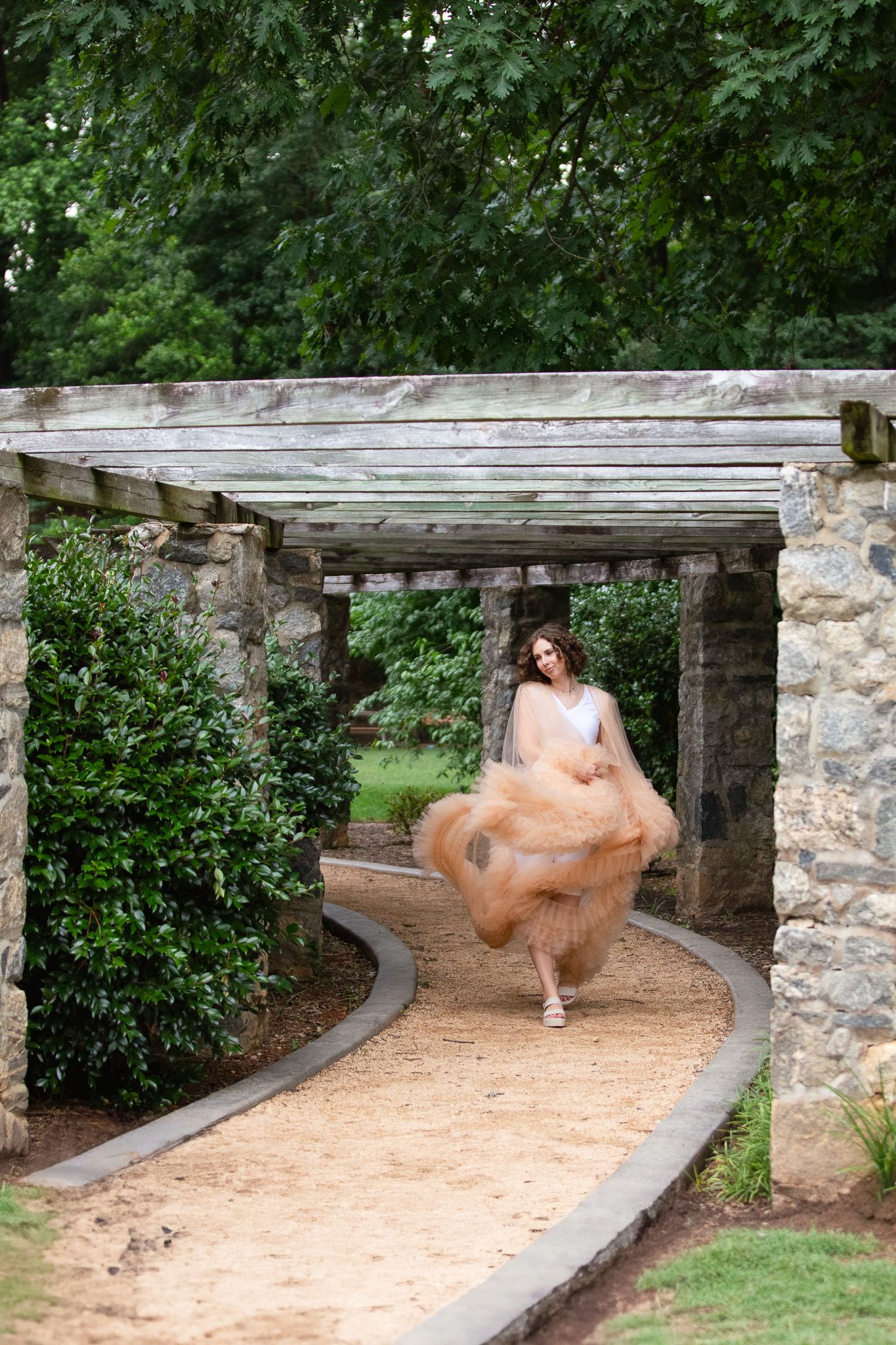 A flowing peach tulle dress spins gracefully along a curved stone path beneath a rustic wooden pergola in a garden setting.