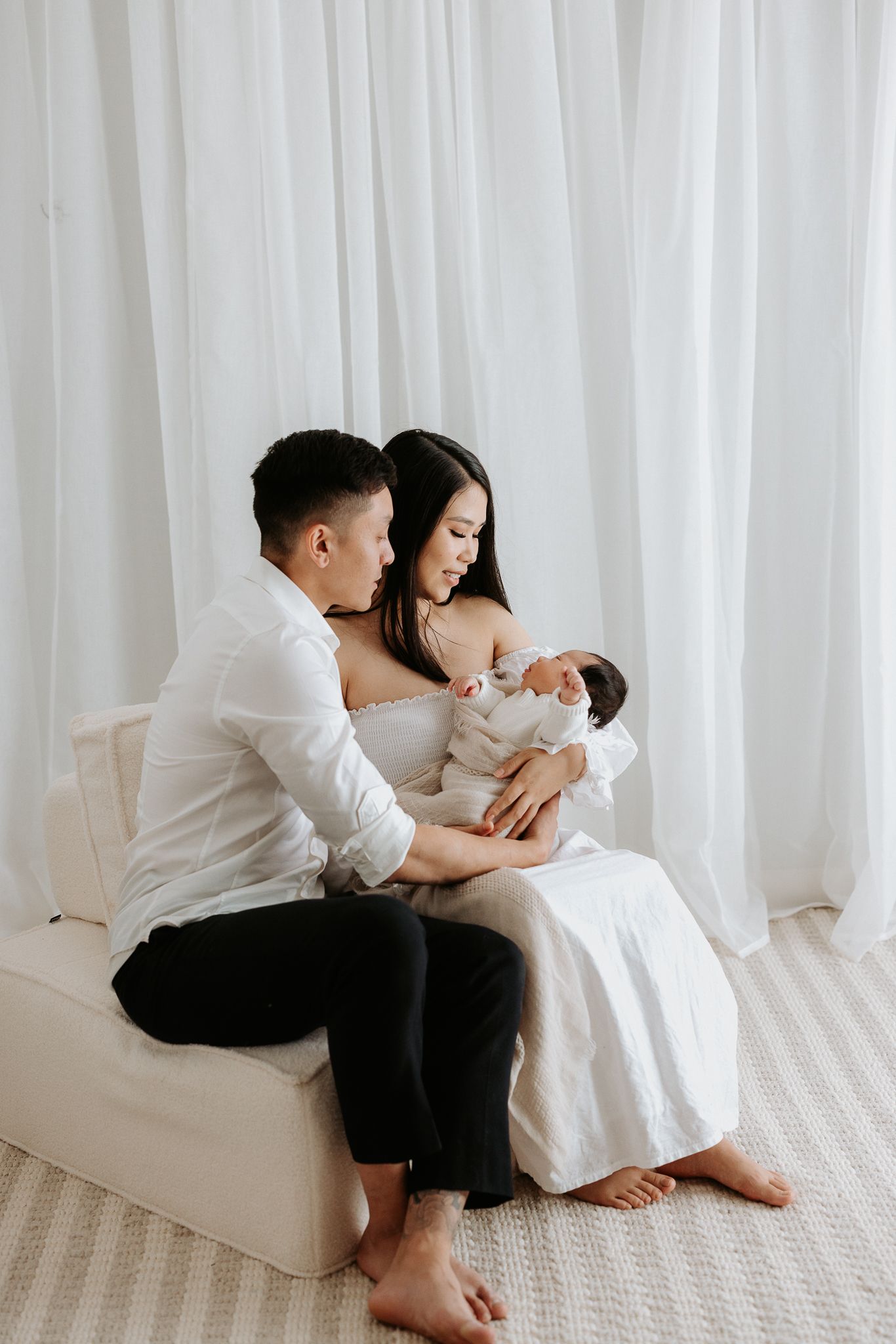 New parents share an intimate moment with their newborn baby in a bright white minimal studio setting.