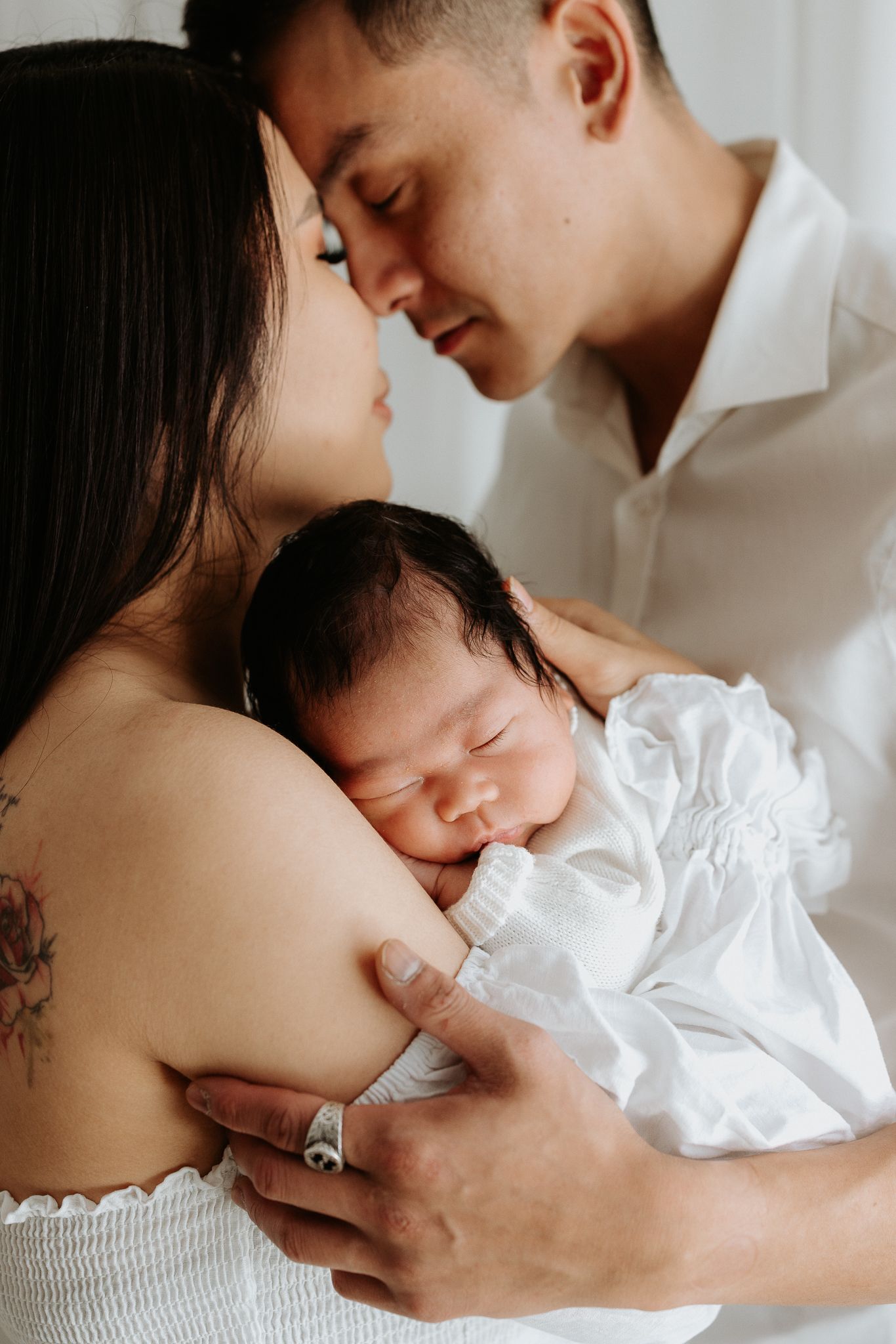 Tender family moment as parents share intimate connection while holding sleeping newborn baby in white dress.