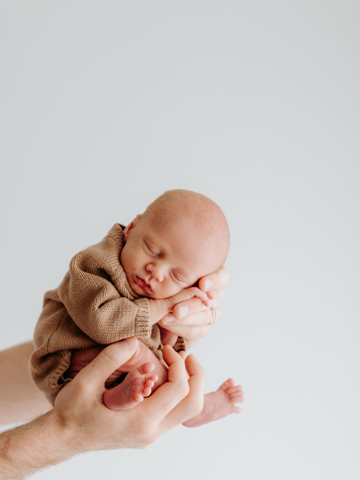 Sleeping newborn baby in brown sweater cradled gently in hands against white background.