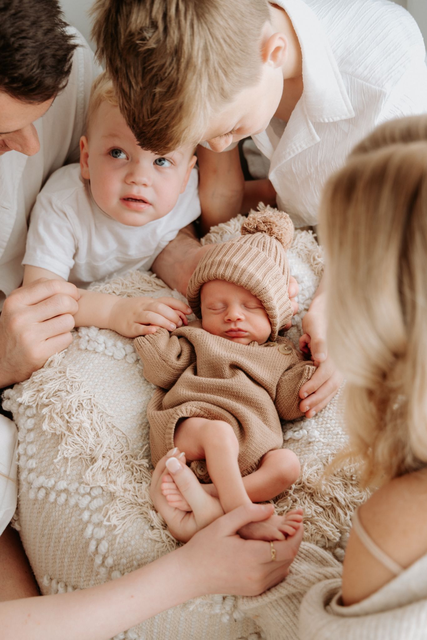 Family gathers around sleeping newborn baby wearing knit hat on textured blanket.