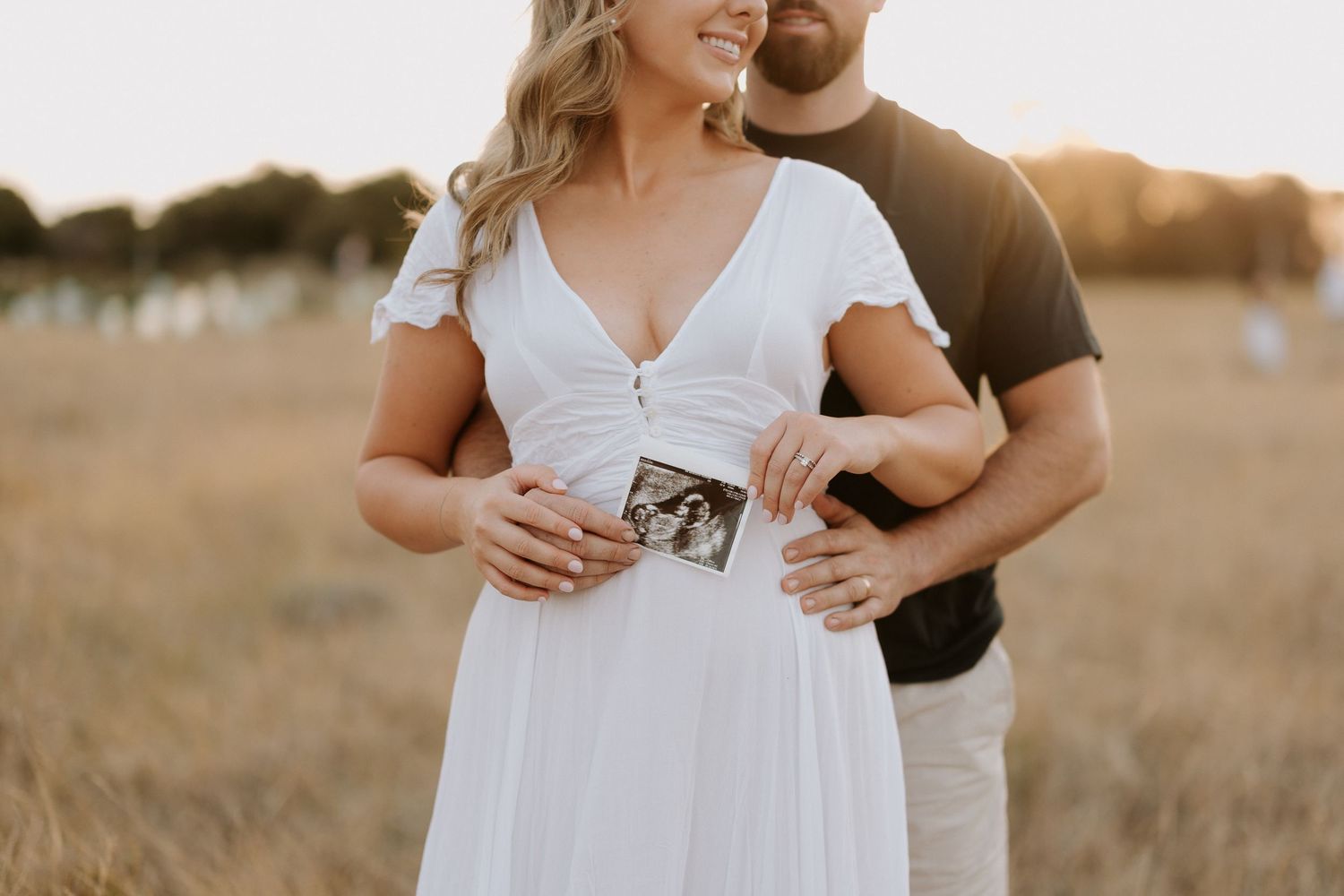 Couple embraces in golden field during sunset while wearing white dress and casual attire.
