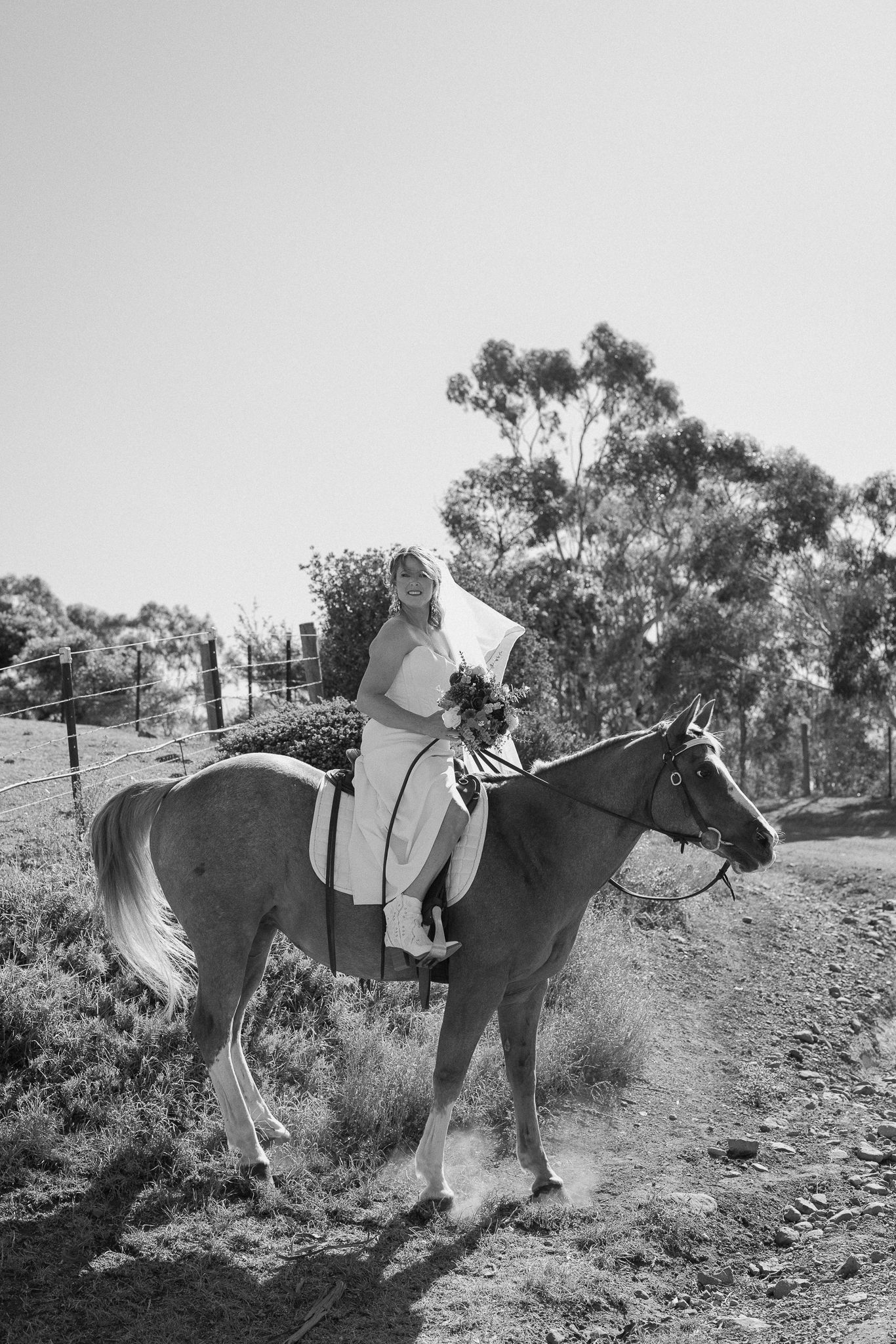 A black and white photograph shows a rider in light-colored attire mounted on horseback against a rural hillside background.