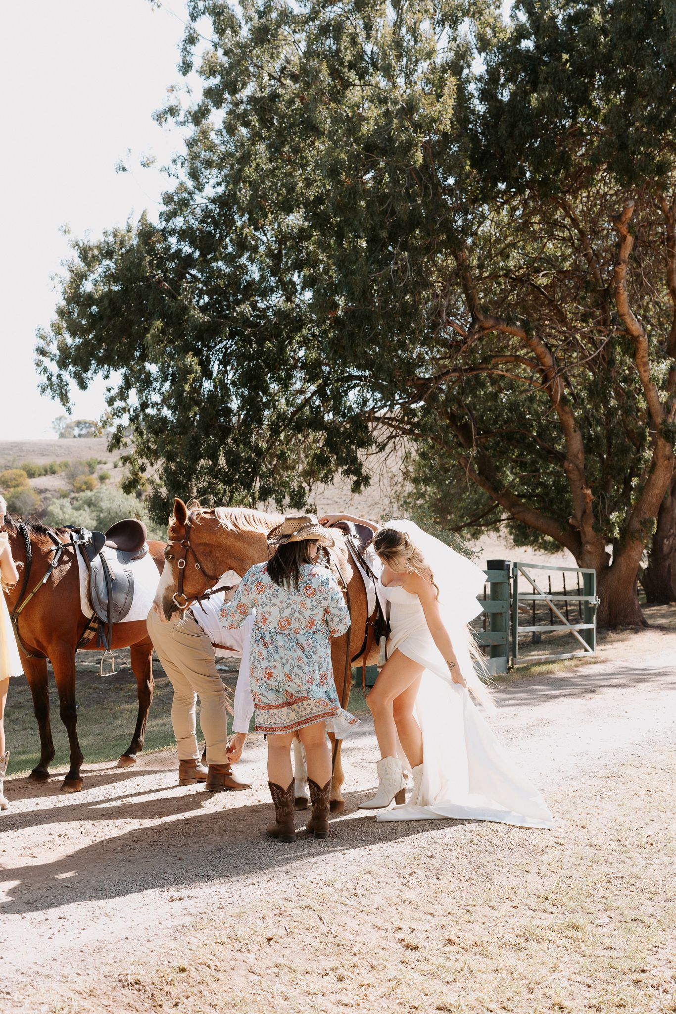 A group gathers around and adjusts their decorative horses on a sunny rural path beneath shaded trees.