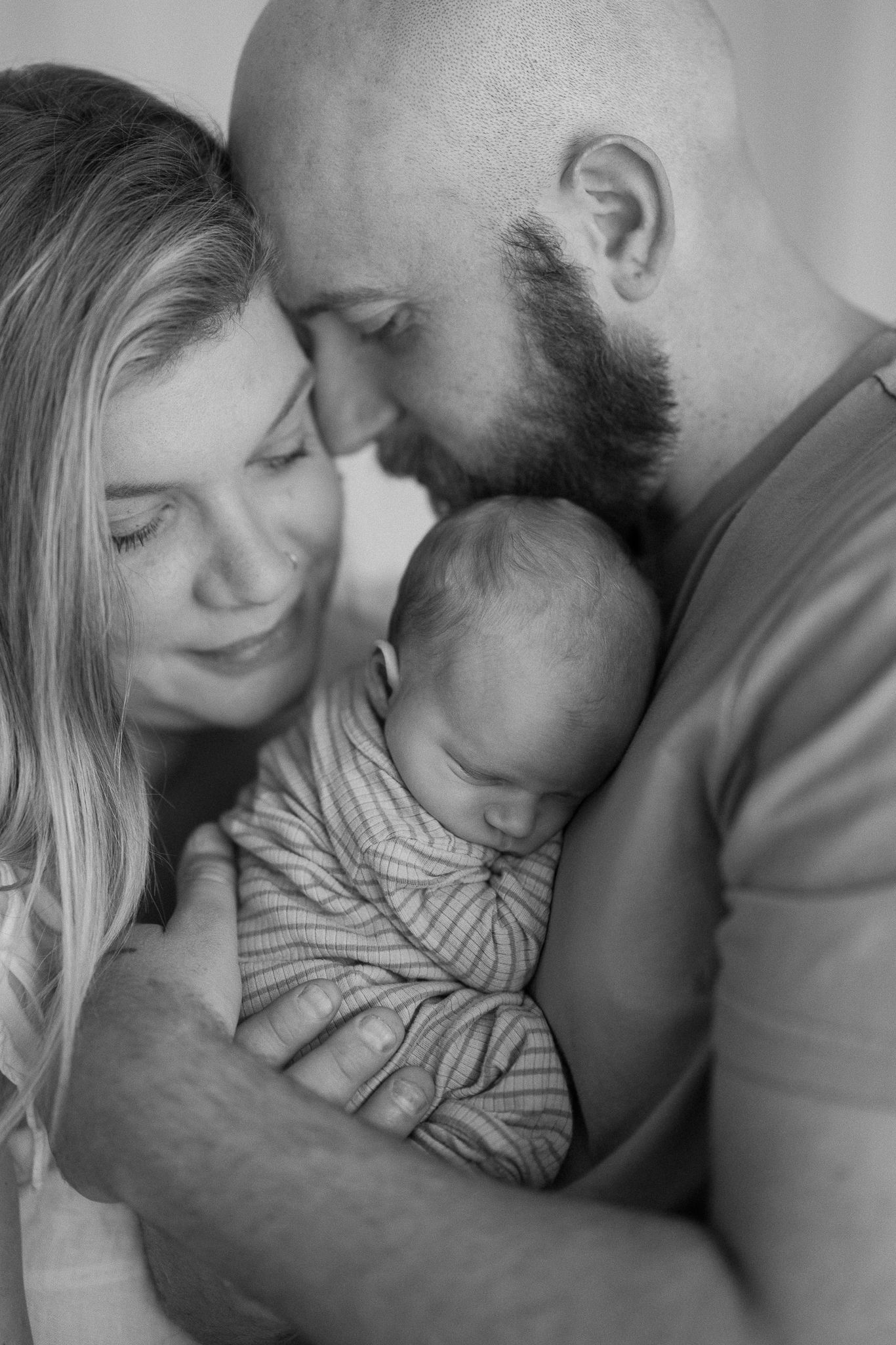 A tender black and white family portrait shows loving parents embracing their sleeping newborn baby.