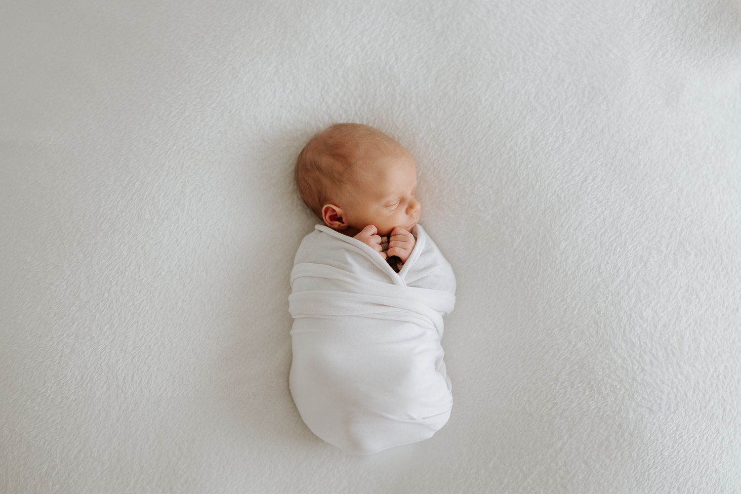 A newborn baby peacefully sleeps while swaddled in soft white blanket on white background.