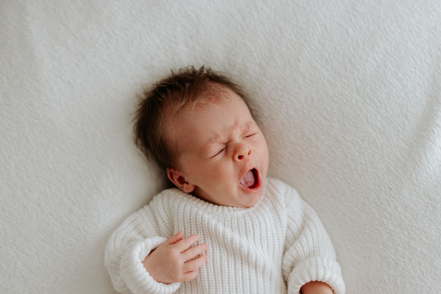 A yawning baby wearing a white knit sweater lies on a soft white blanket looking sleepy.