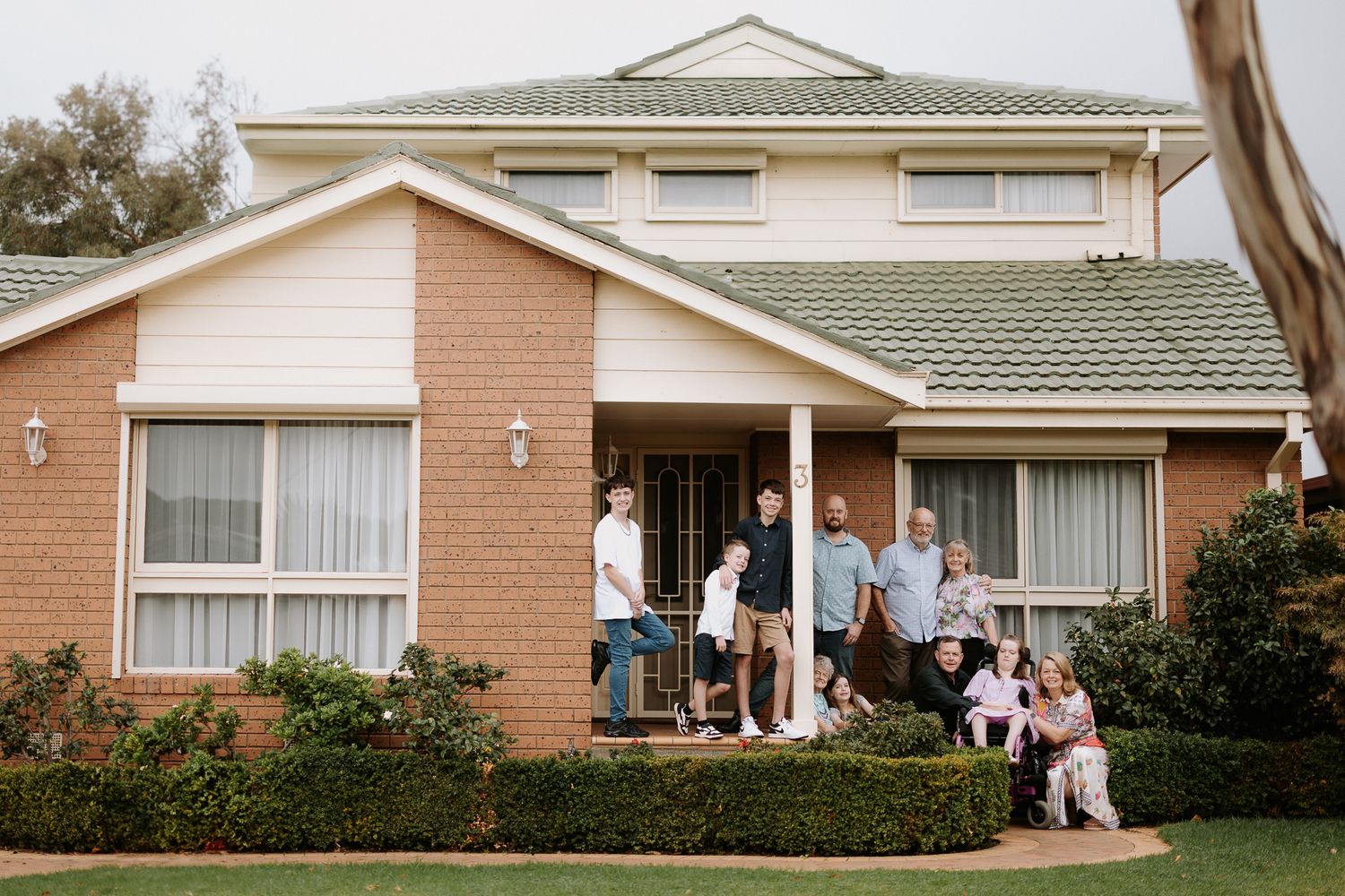 A group stands together on the front porch of a pink brick two-story suburban home with a green roof.