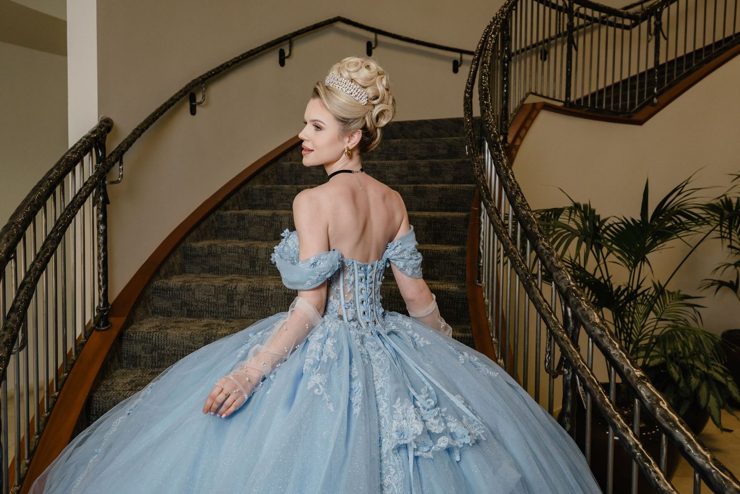 A model in a light blue off-shoulder ballgown with a corseted bodice poses on an elegant curved staircase.