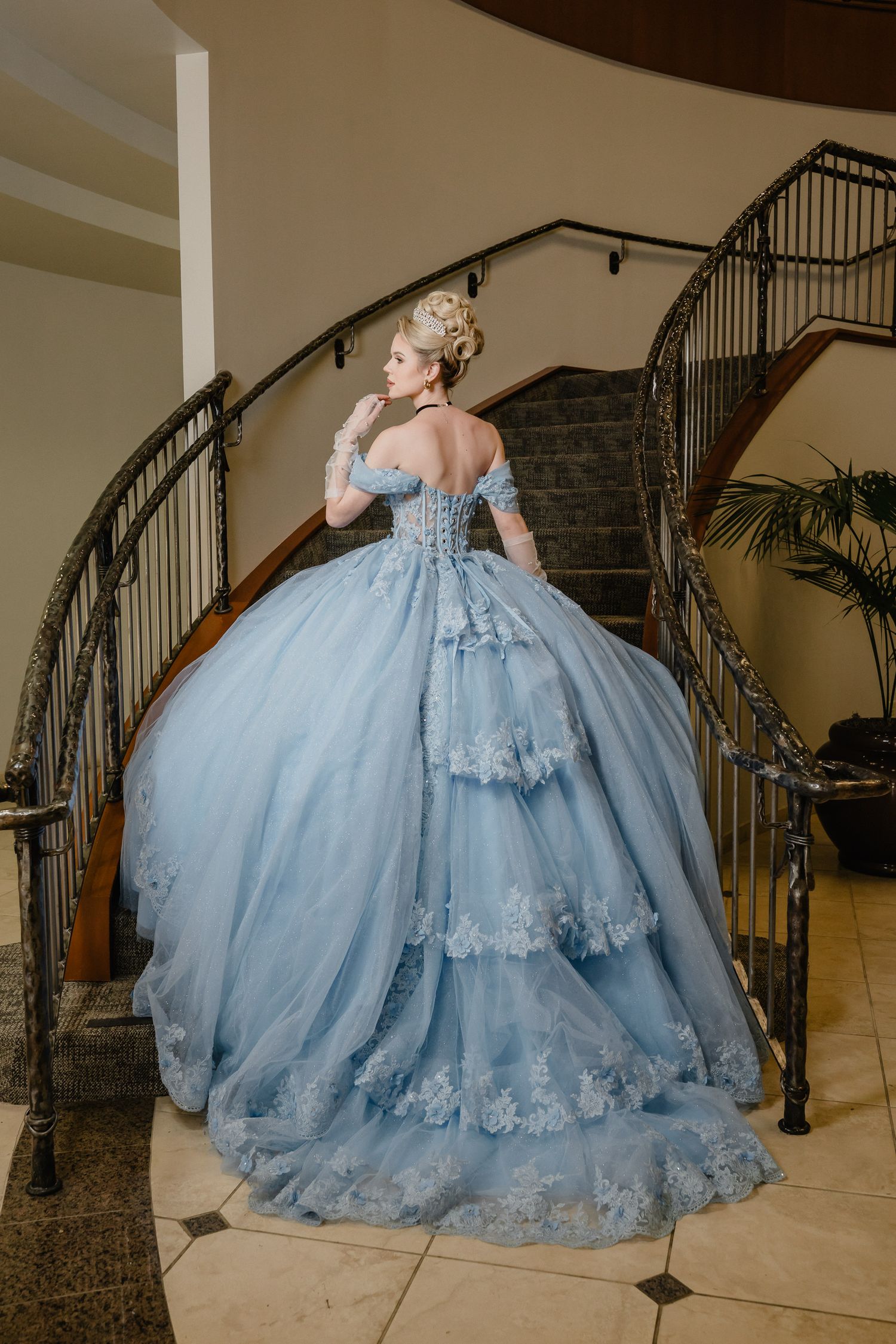 Elegant light blue ball gown with off-shoulder design and tiered lace details shown on grand staircase with wrought iron railing.