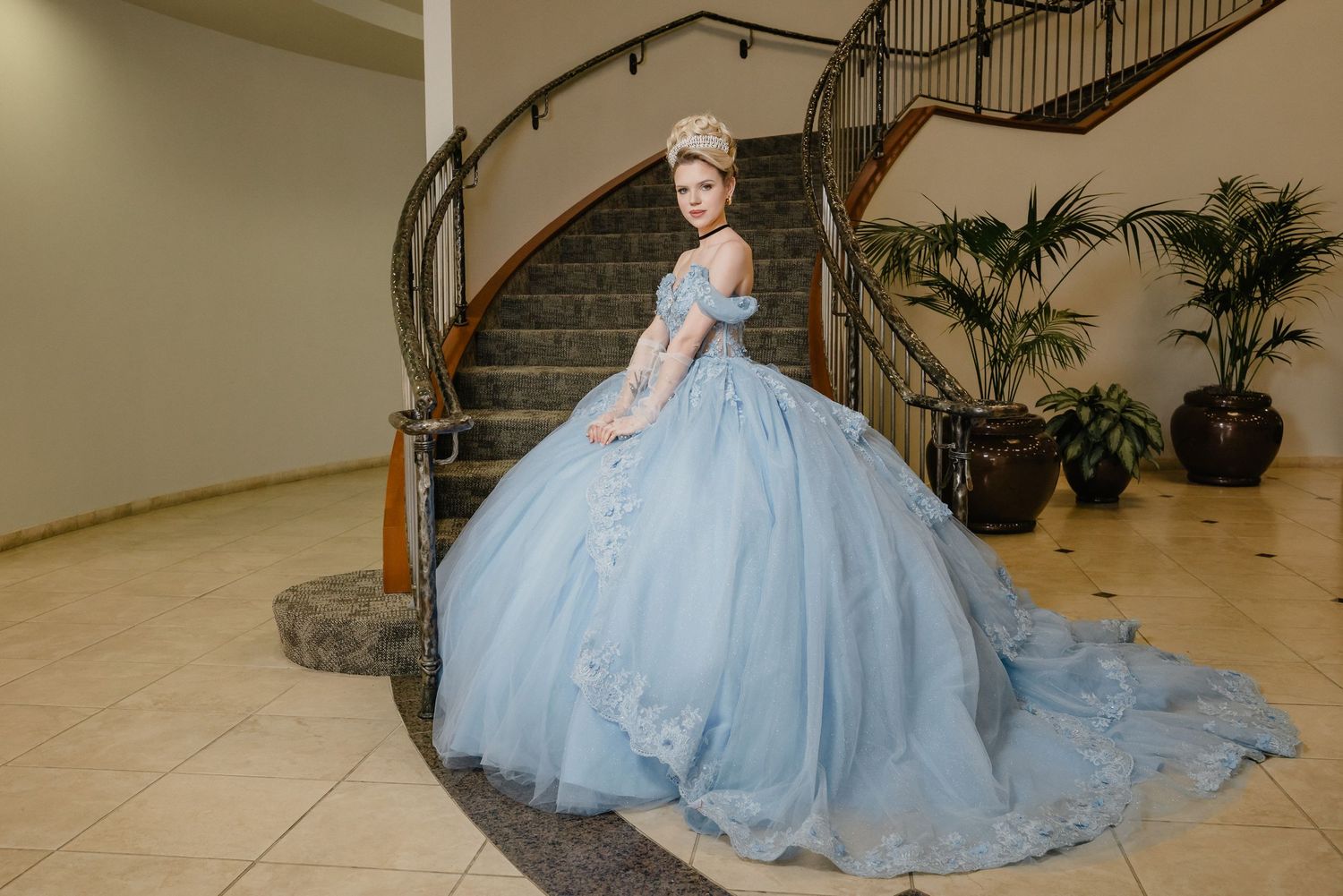 An elegant powder blue ballgown with full skirt and long train photographed against a curved staircase and potted palms.