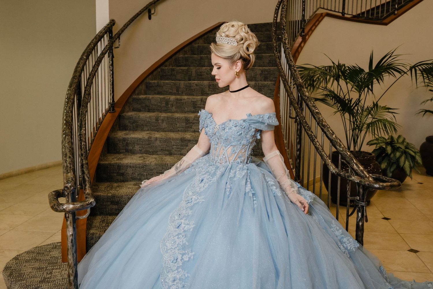 Beautiful light blue ball gown with off-shoulder design and floral appliques stands elegantly by a curved staircase. Cinderella