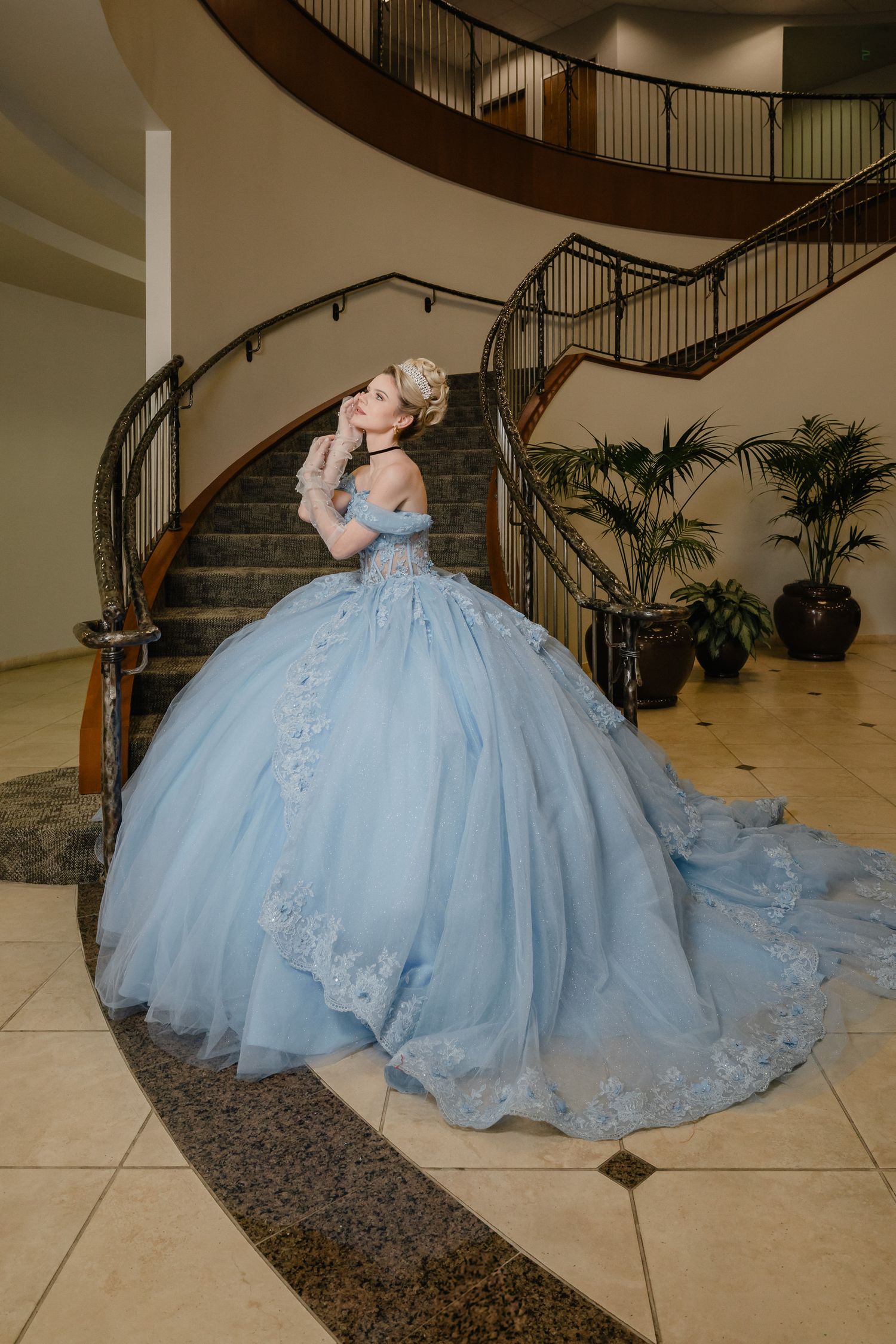 A light blue ballgown with floral lace details and a voluminous tulle skirt flows elegantly on a curved staircase.