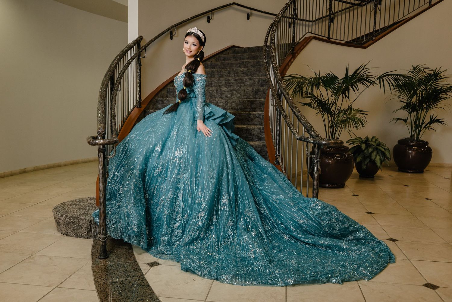 A person in a stunning turquoise blue ball gown with glittering details poses on an elegant spiral staircase.