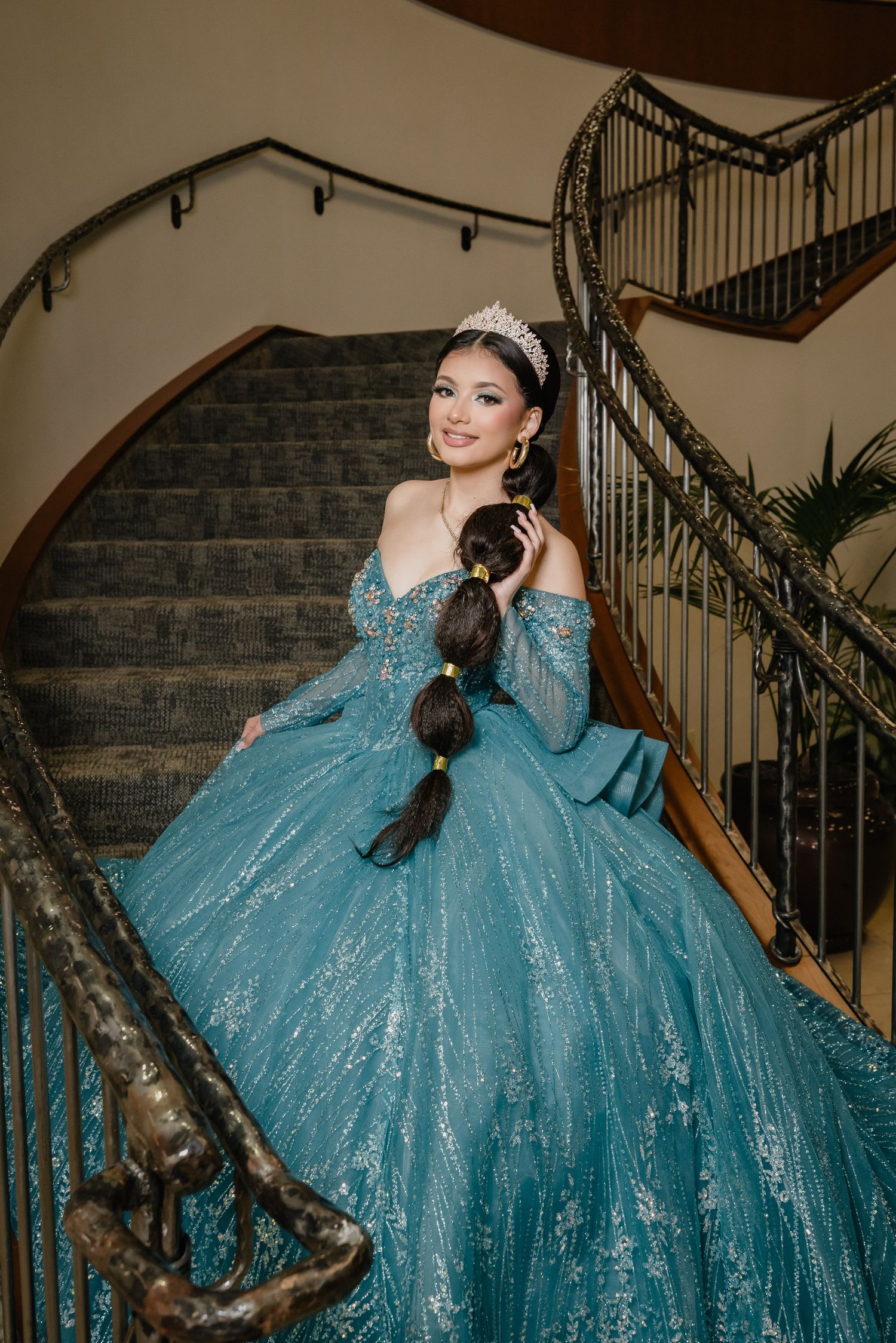 A person in a sparkling turquoise ball gown poses on an elegant curved staircase with wrought iron railings.