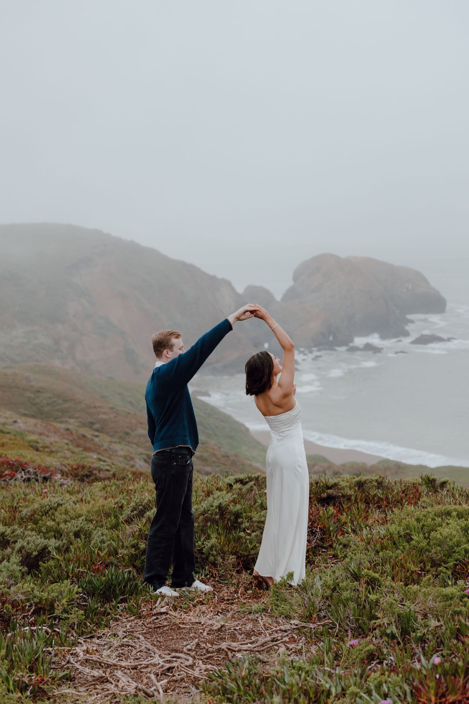 A romantic couple embraces on a misty beach with rocky cliffs and crashing waves in the background at sunset.