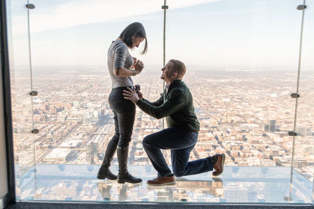 A marriage proposal takes place on a glass observation deck high above a sprawling city landscape during daytime.