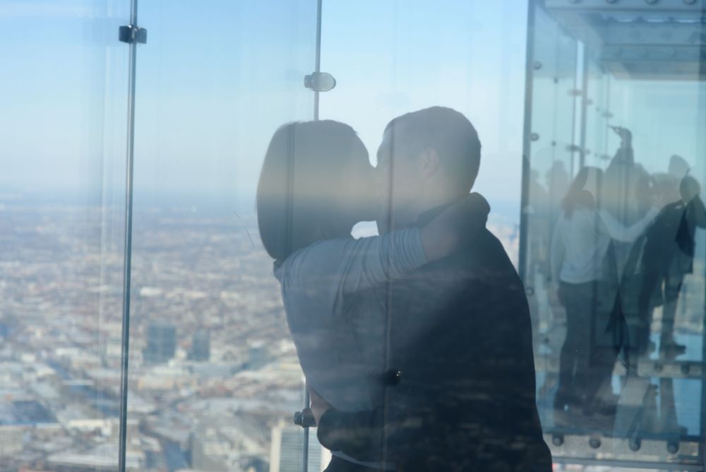 Silhouetted figure looking out over cityscape from high-rise glass viewing platform on clear day.