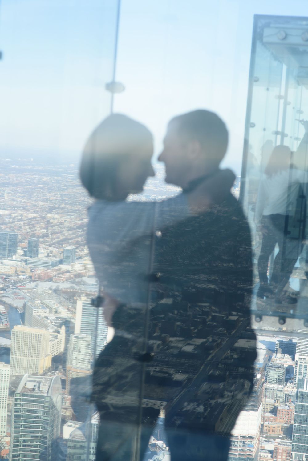 A couple stands close together against a reflective glass window overlooking a vast cityscape from a high vantage point.