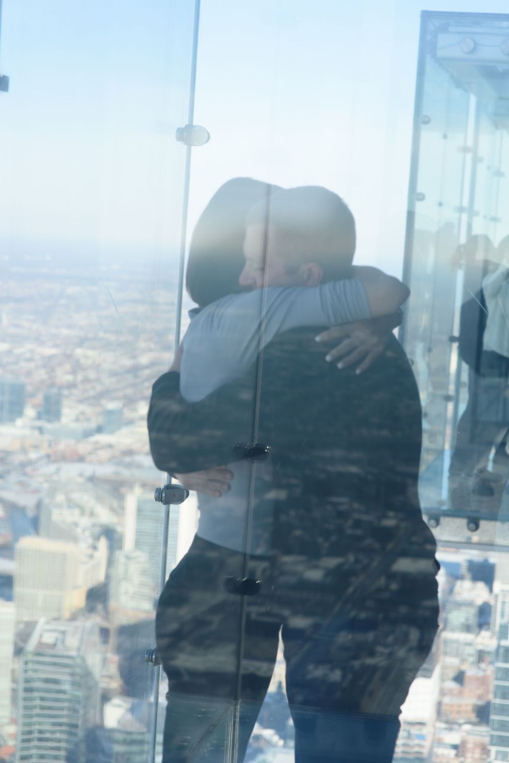 Two people share an embrace while standing in a glass observation deck high above a cityscape on a hazy day.