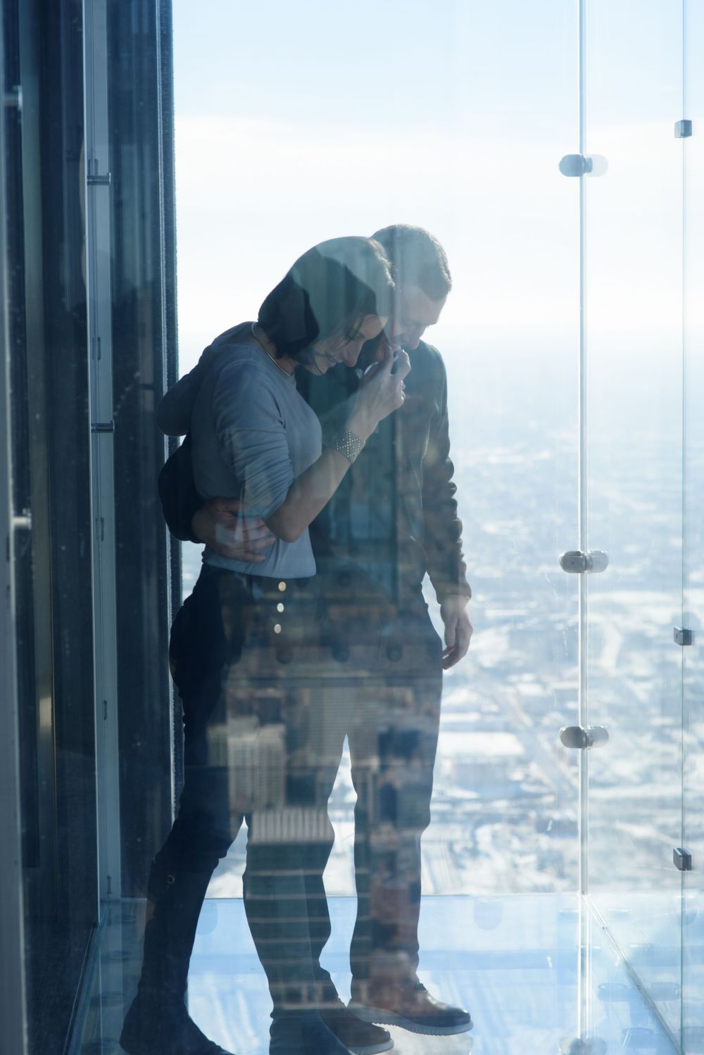 A couple stands close together in an observation deck or glass walkway high above a cityscape on a sunny day.