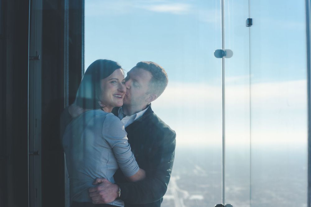 A couple embraces while looking out over a city view from a glass-enclosed observation deck or tall building.