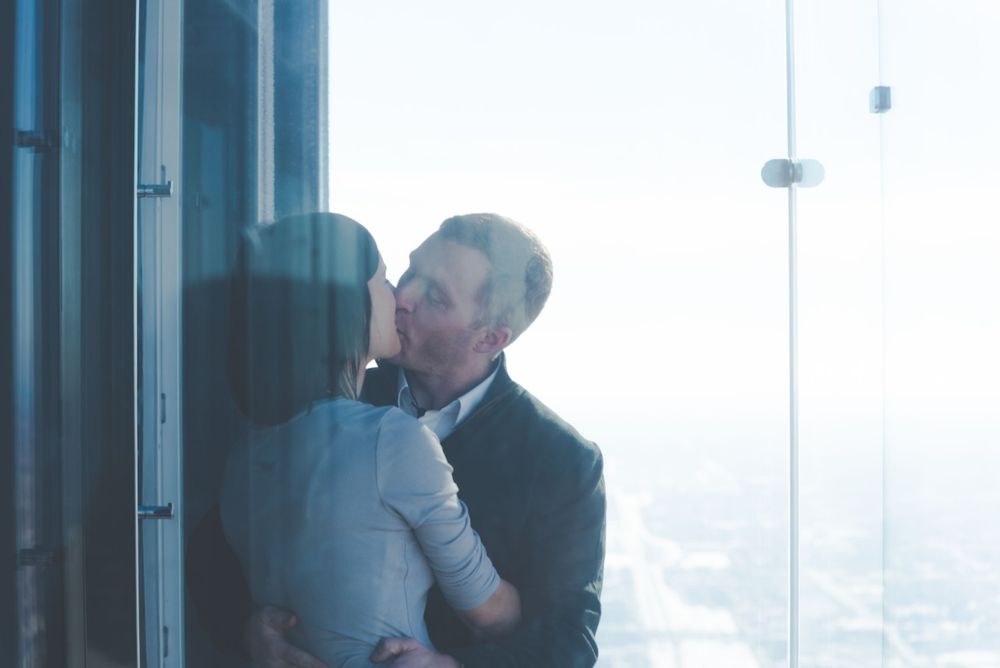 A couple shares a romantic kiss by large windows in a high-rise building with a misty cityscape view in the background.