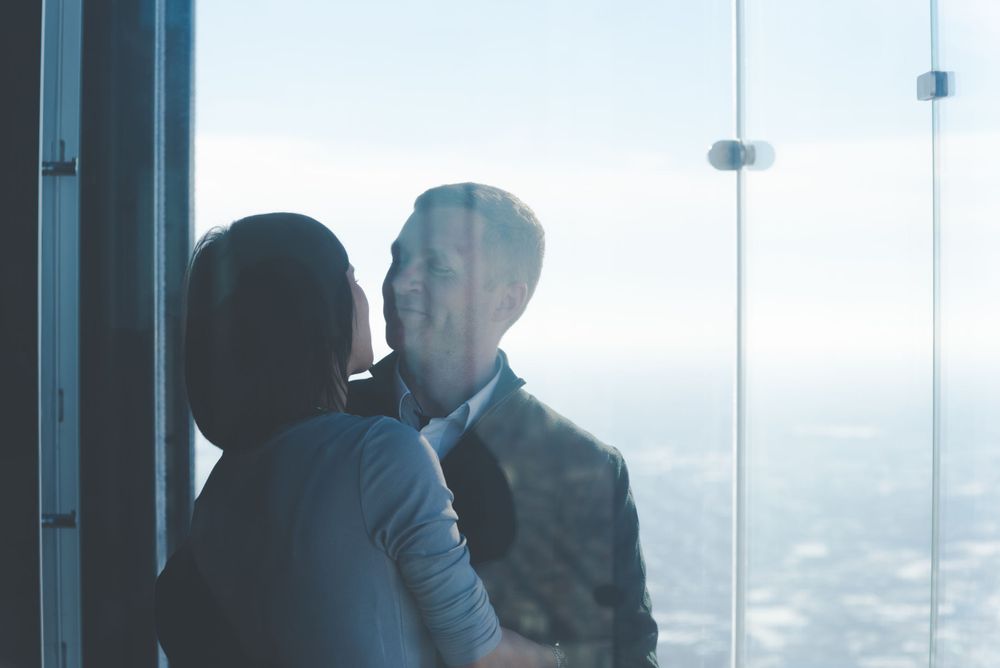 Two silhouetted figures share an intimate moment against a bright window overlooking a cityscape.
