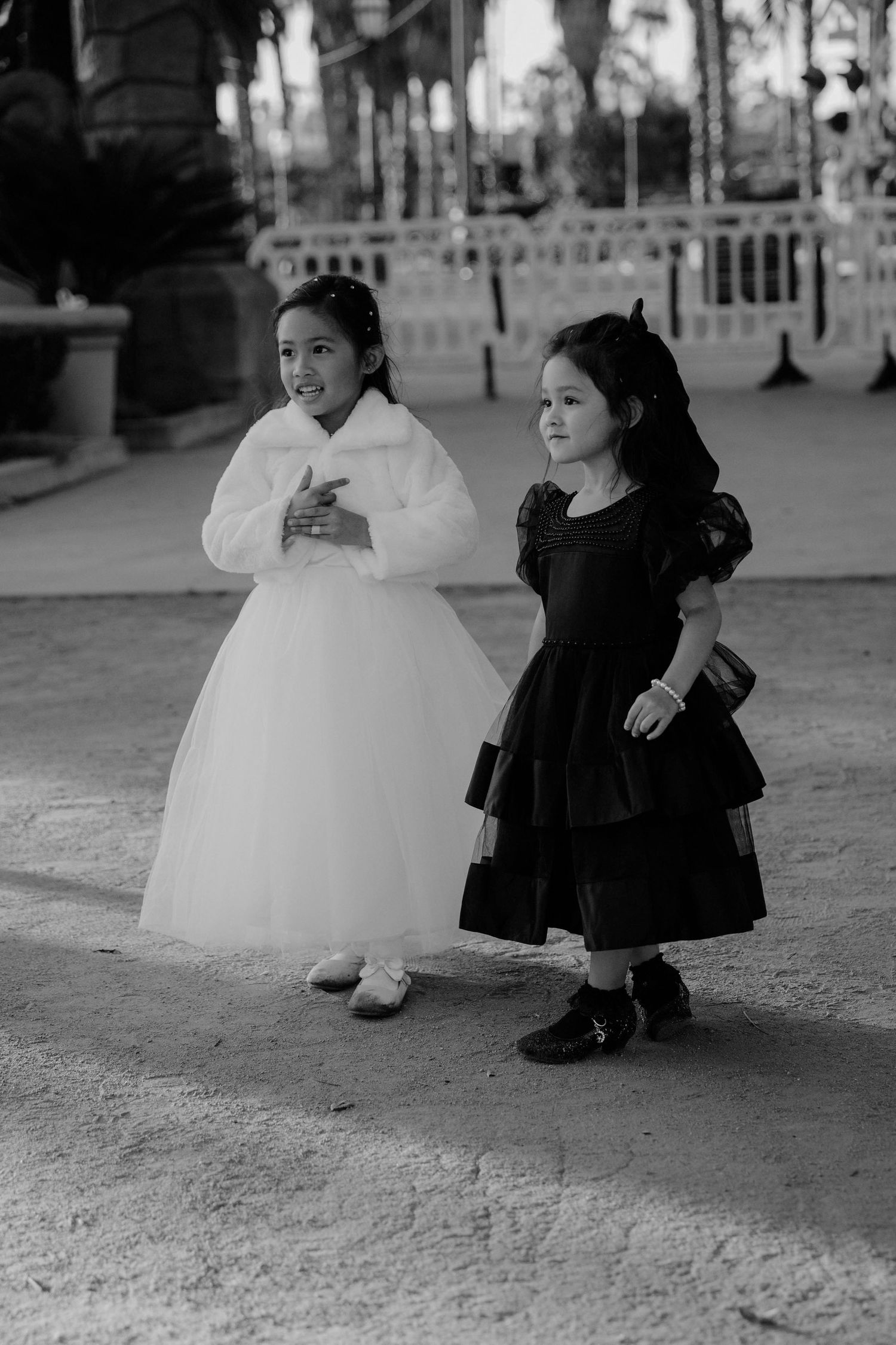 Black and white photo of two young children in formal dresses standing outdoors, one in white and one in black.