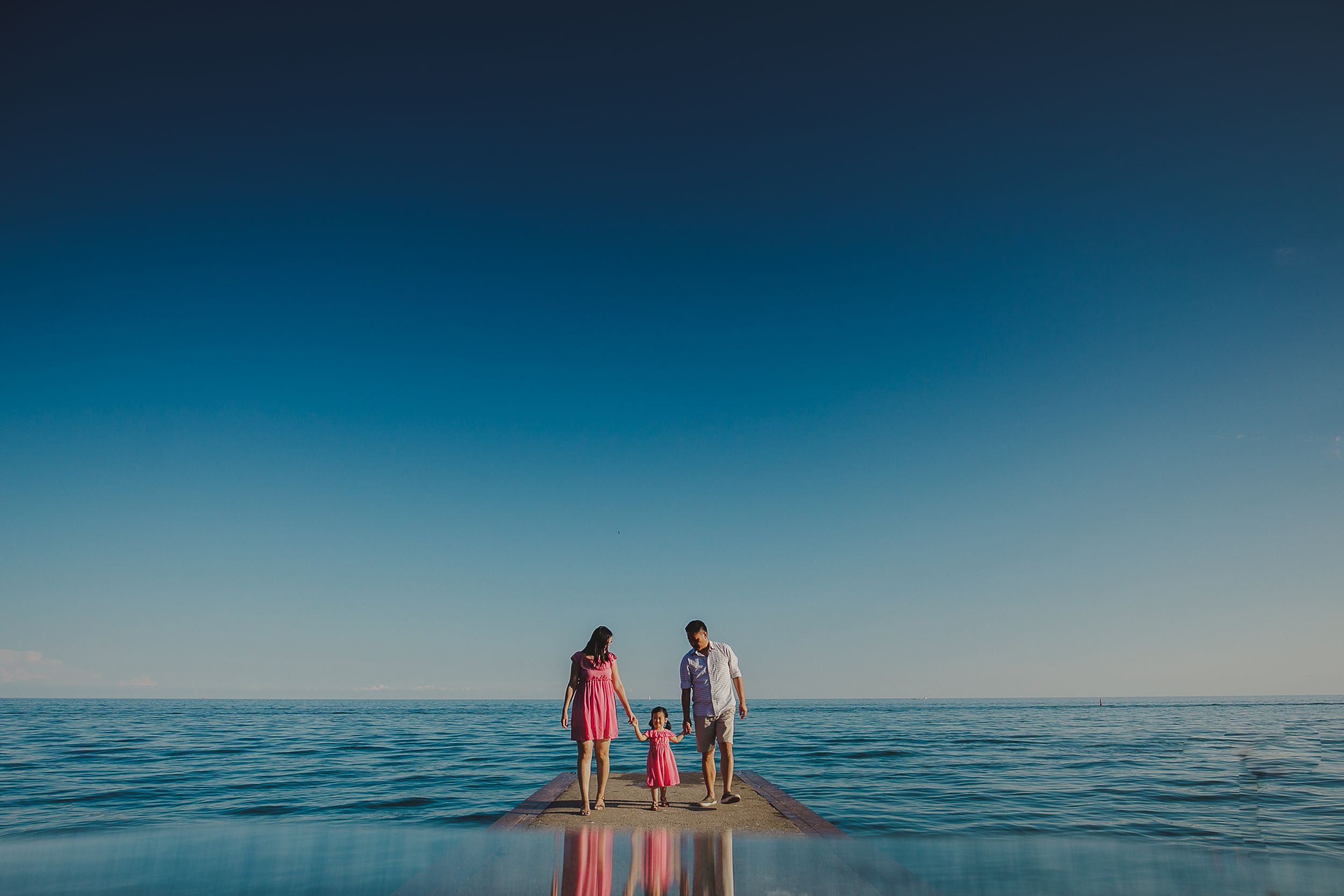 Family holding hands while standing on a wooden pier over calm ocean waters during sunset with blue sky.