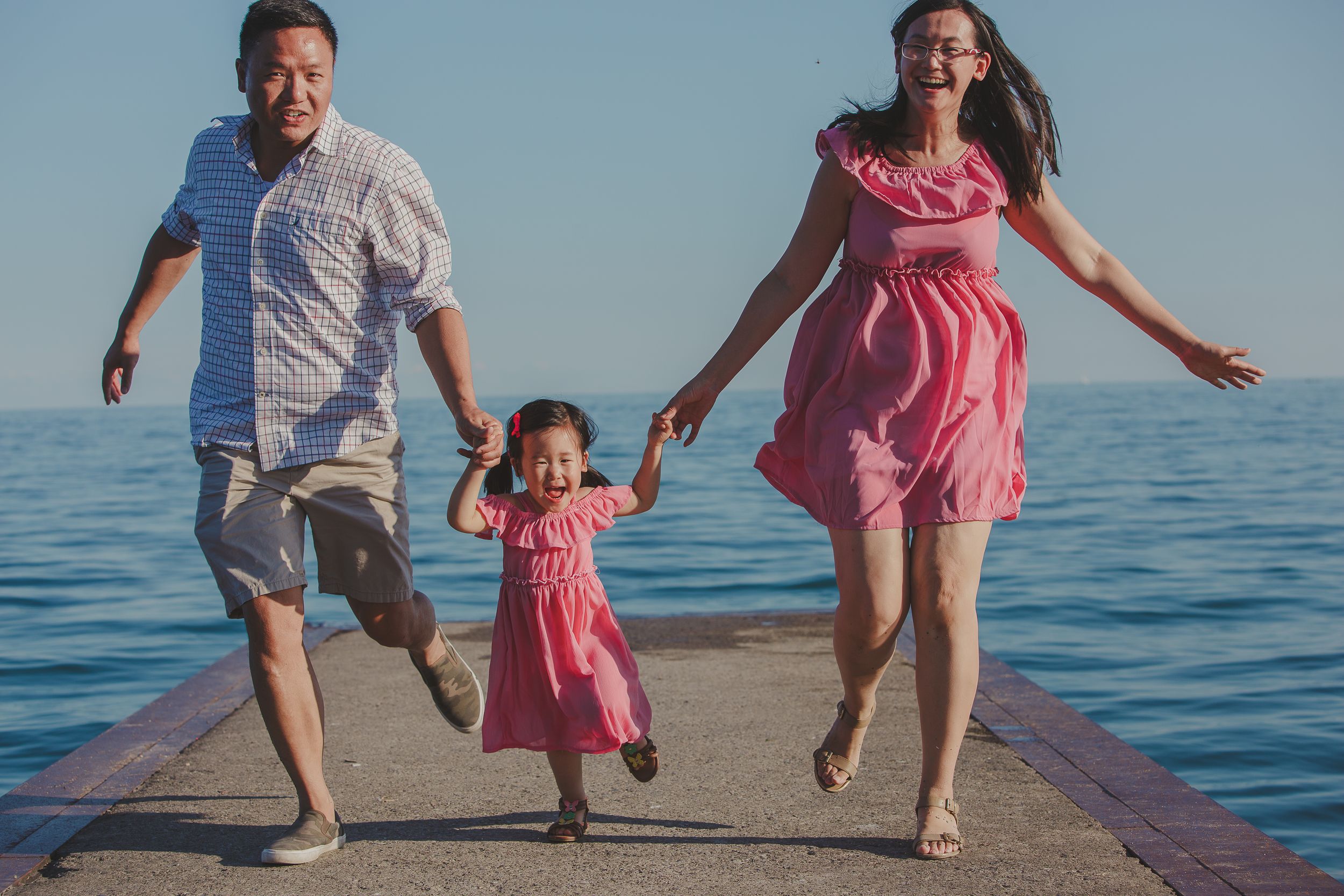 A joyful family leaps together on a wooden pier over calm blue ocean waters on a sunny day.