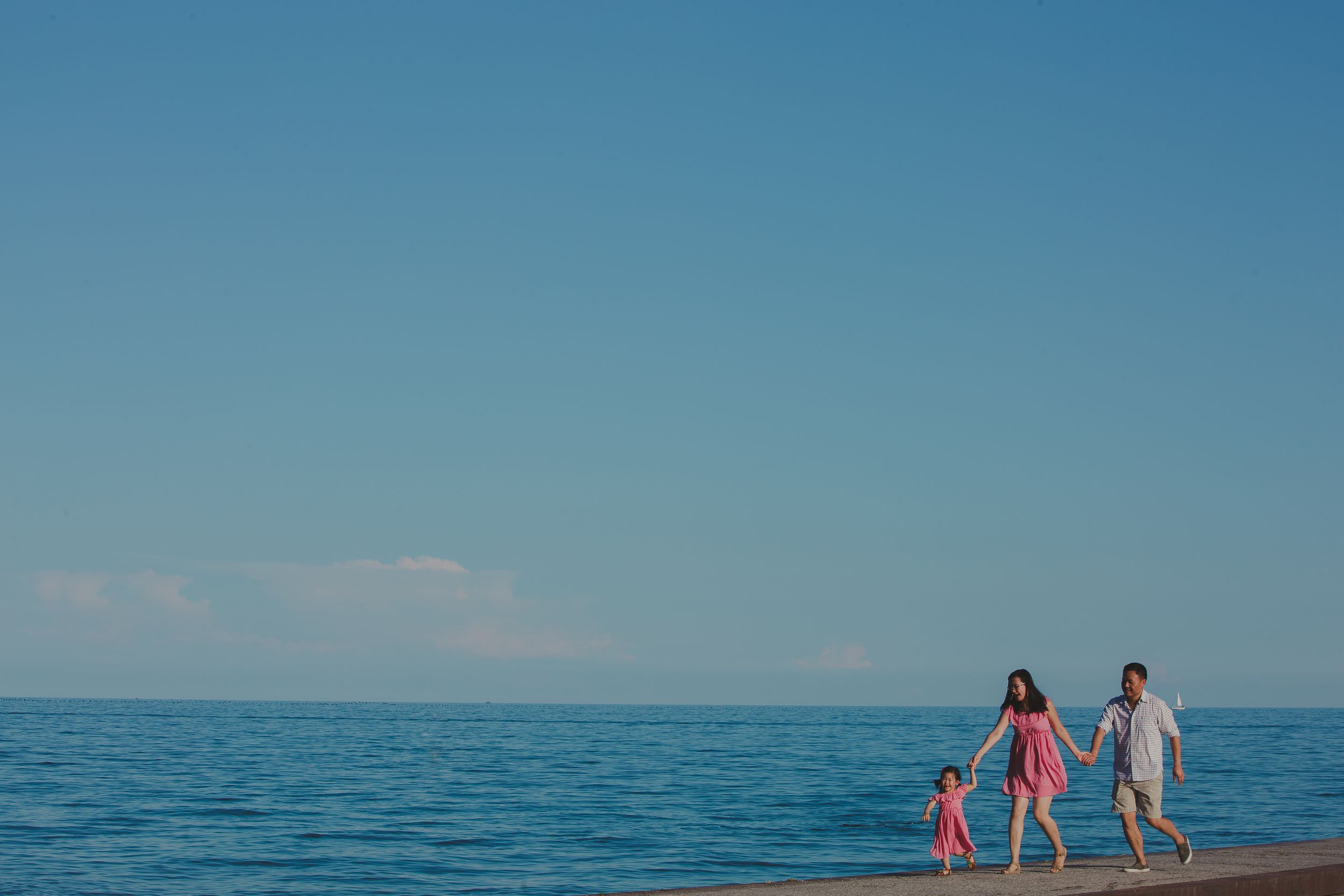 A family holding hands while walking along the shore of a calm blue ocean under clear skies.