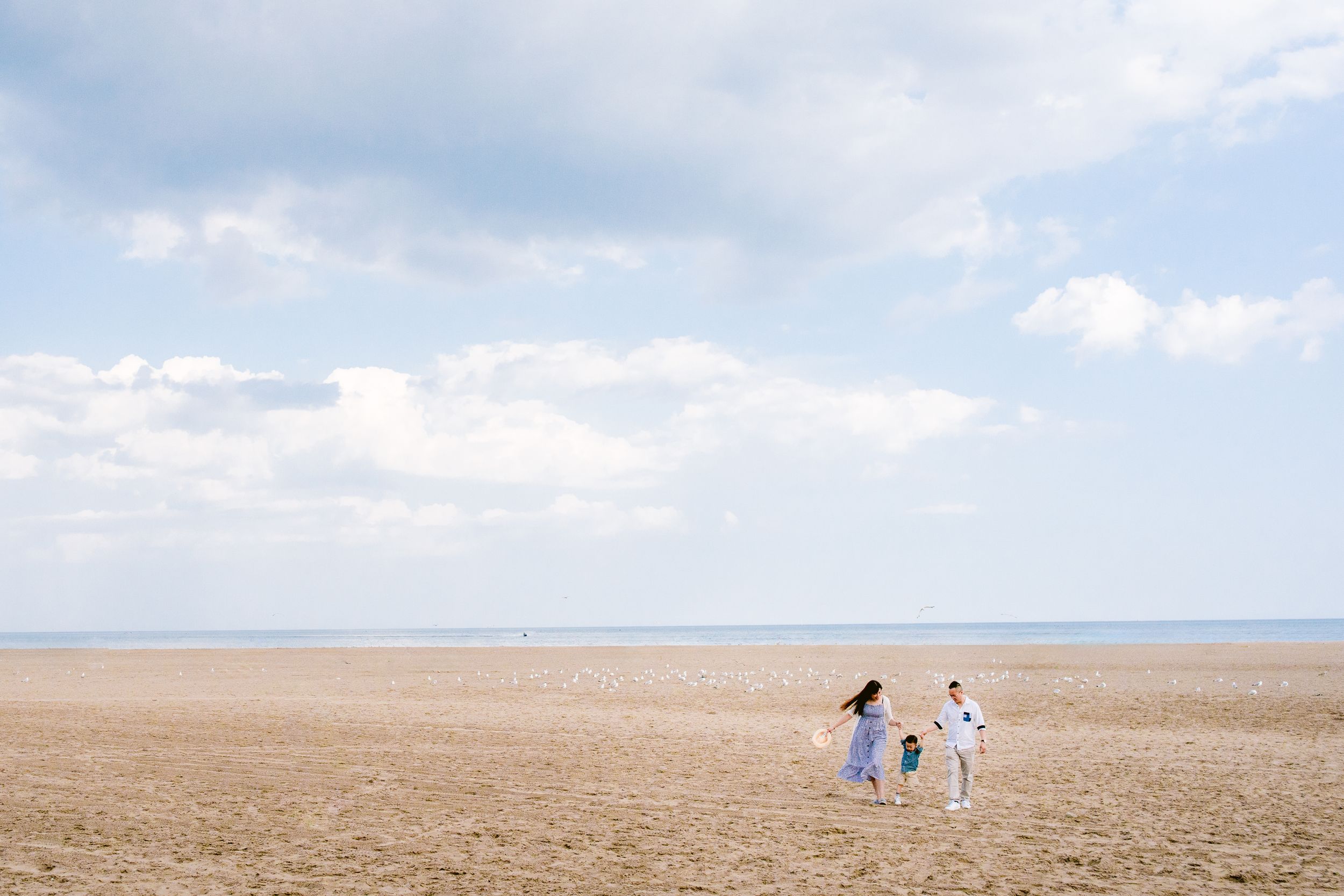 Two people and a child walk across a vast empty sandy beach under a bright blue sky with white clouds.