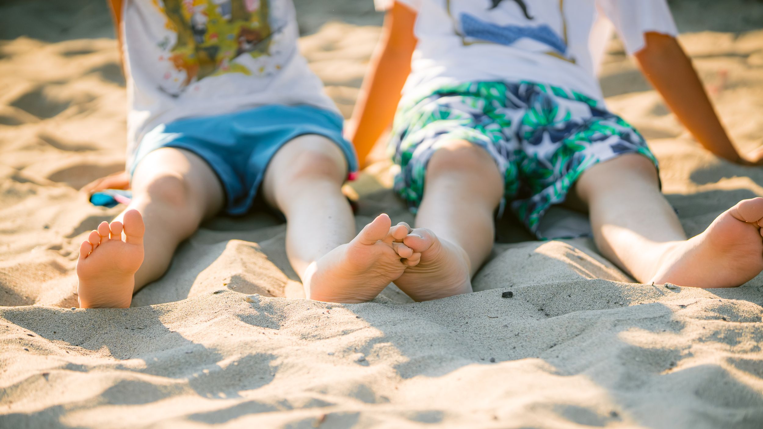 Two sets of bare feet belonging to children laying on warm beach sand in summer weather.