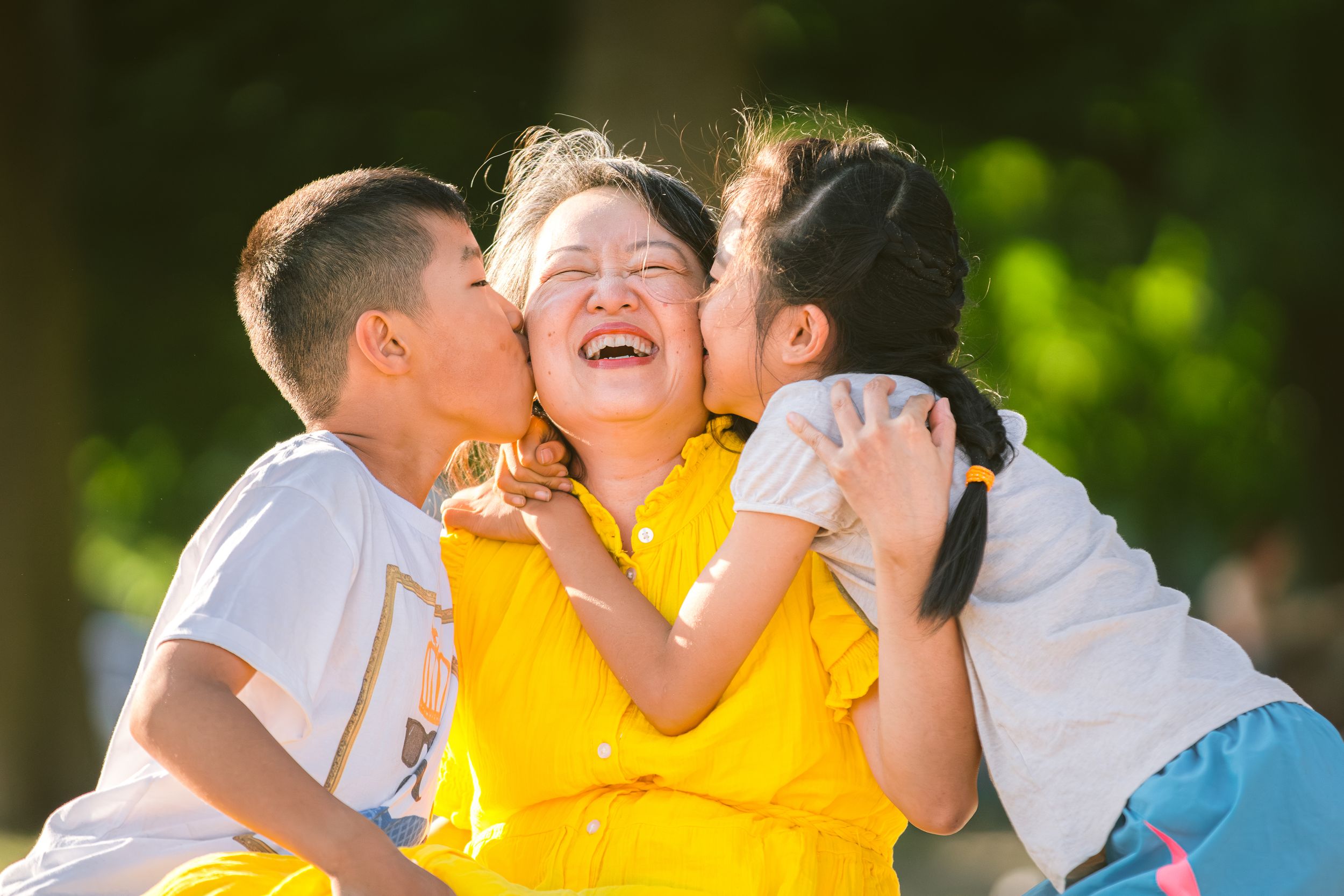 Three joyful family members share an affectionate moment outdoors in bright sunlight.