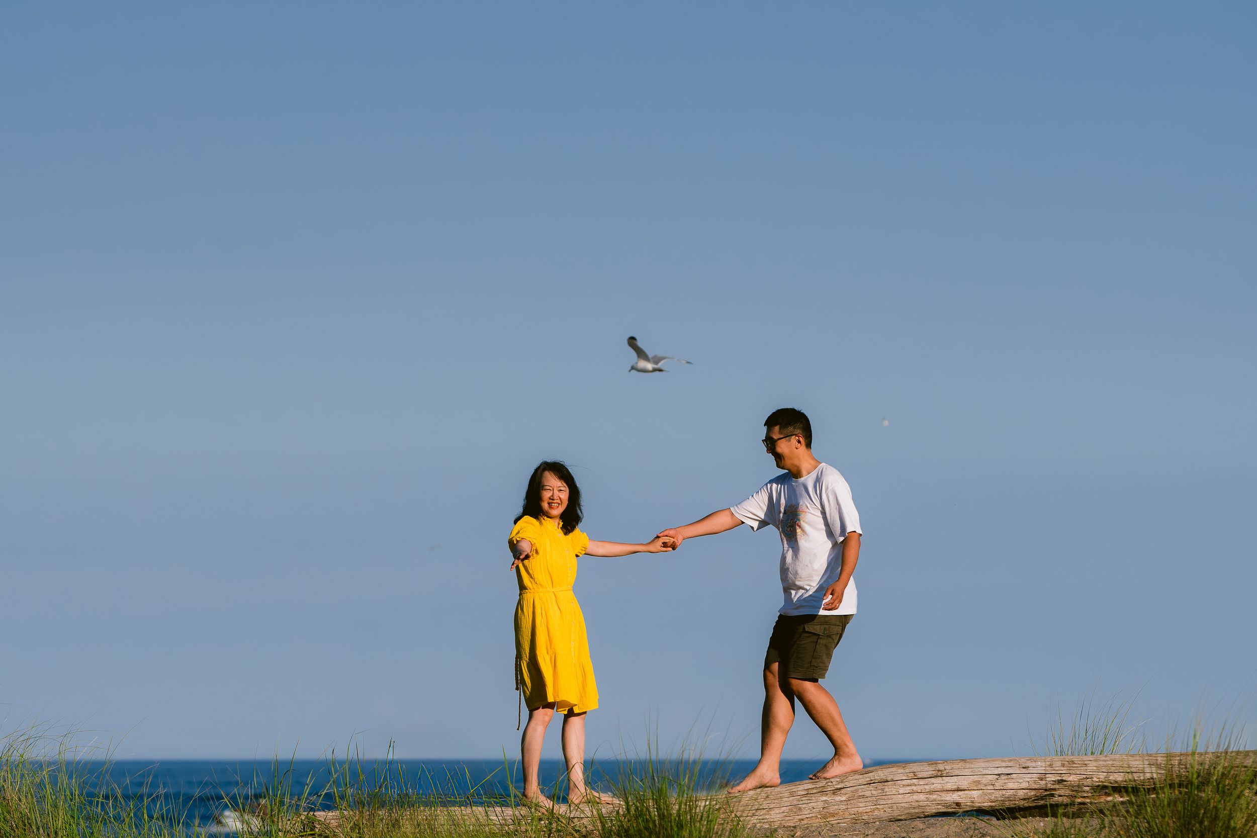 Two people hold hands while standing on a grassy hill against blue sky and ocean background during daytime.