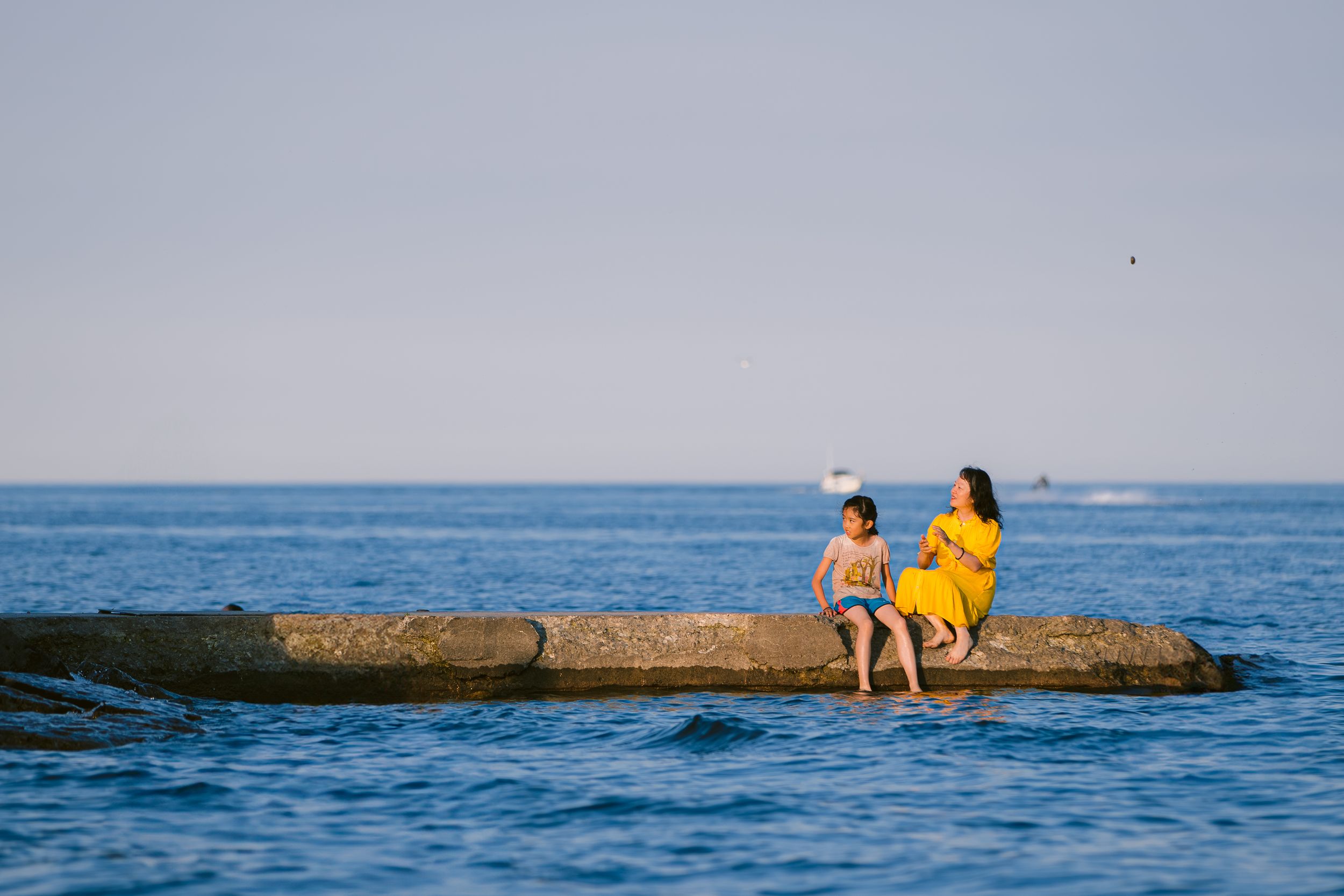 Two people in yellow clothing sit on a rocky outcrop in the ocean on a clear summer day.
