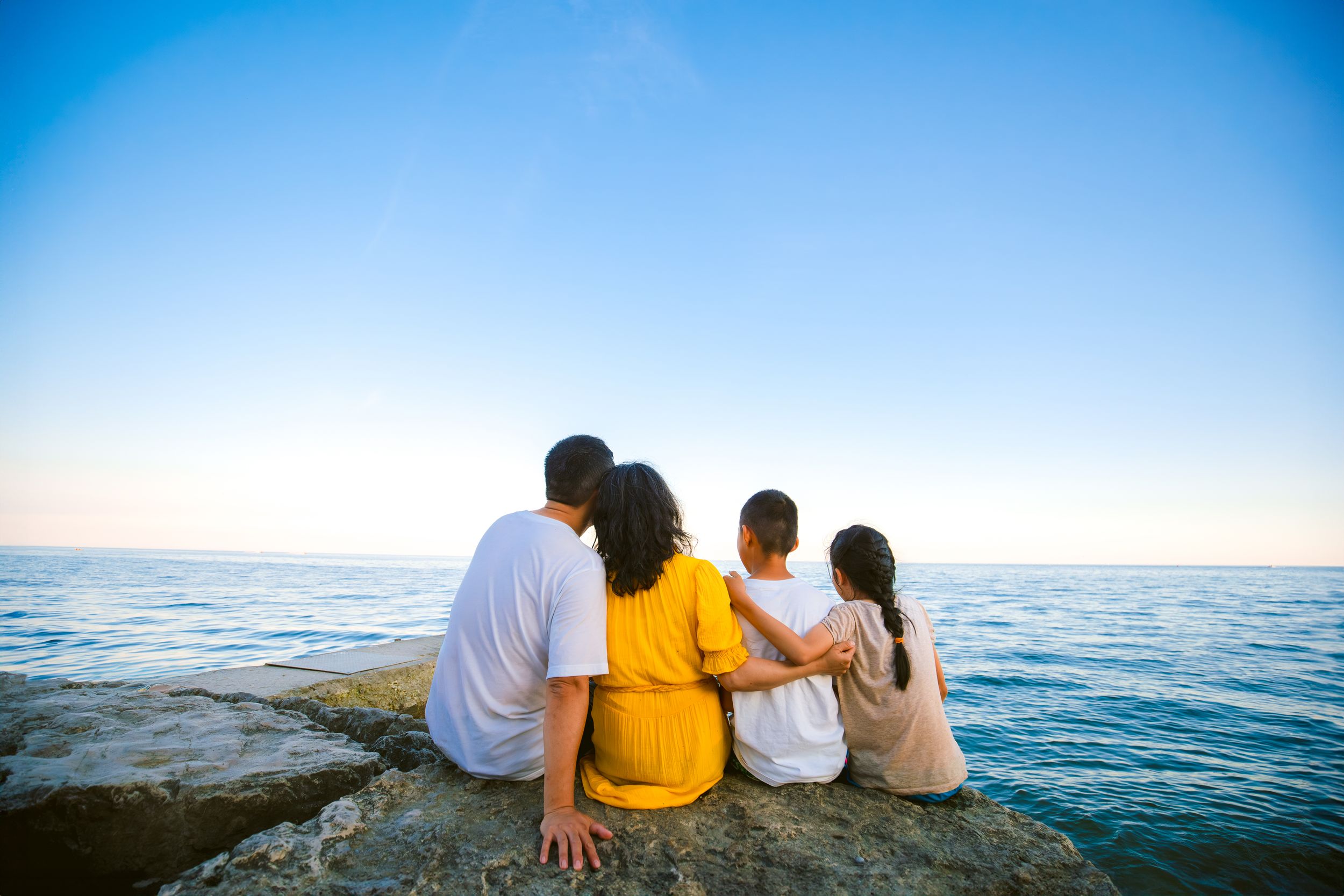 Family of four sits together on rocky shore overlooking calm blue ocean on sunny day.