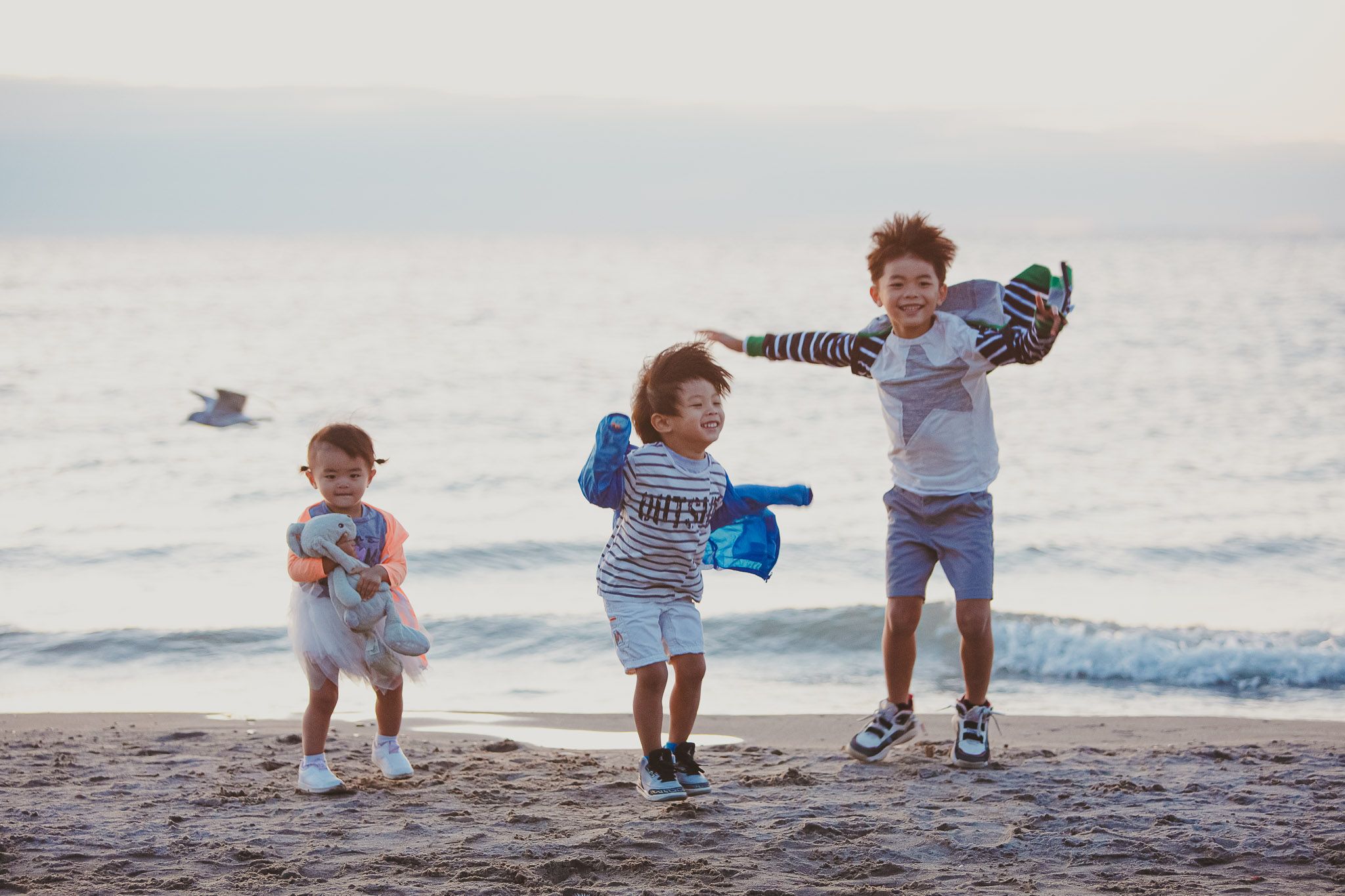 Three joyful children playing and jumping together on a beach at sunset with waves in the background.