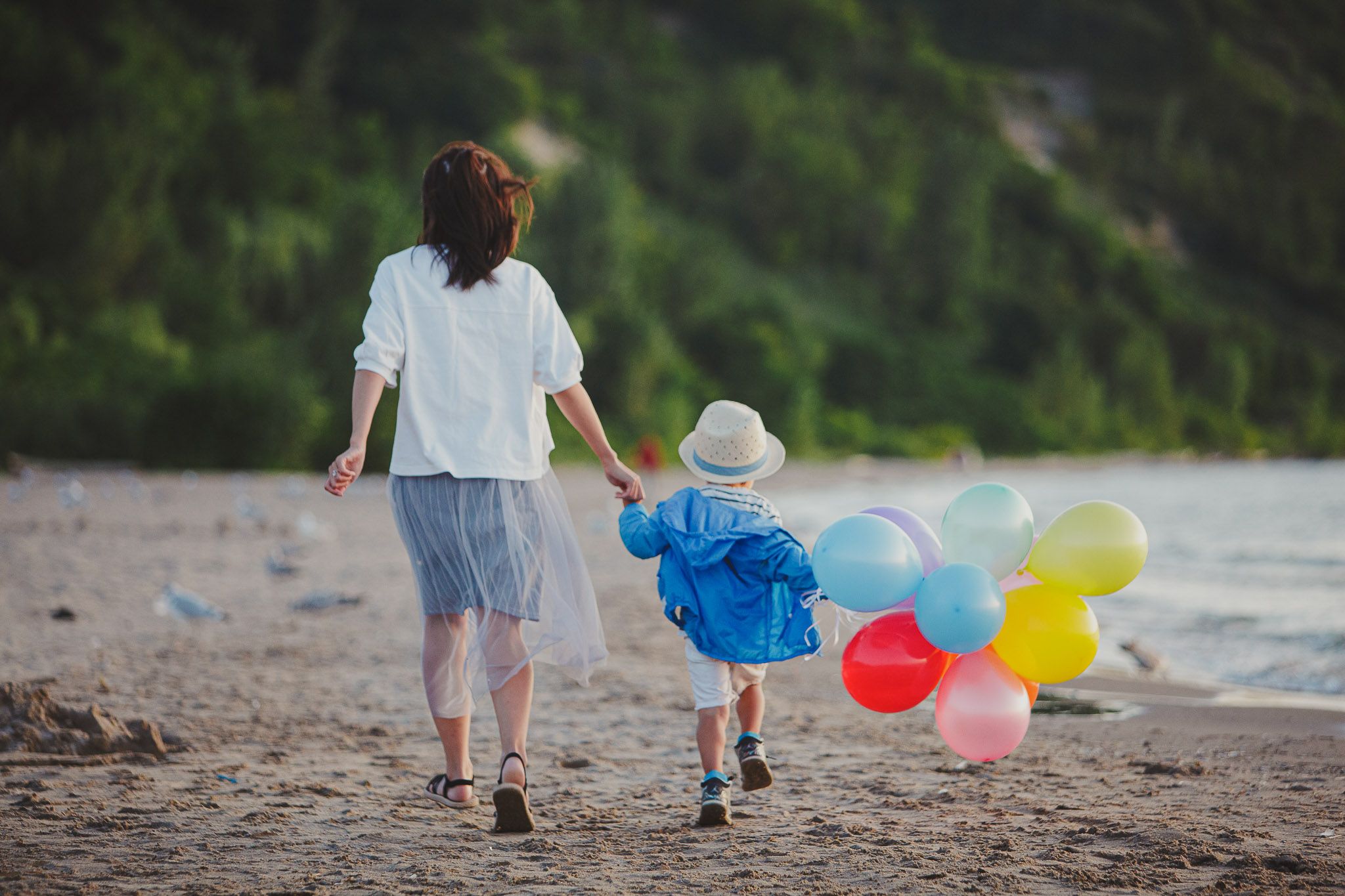 Two figures walk hand in hand along a beach shore while holding colorful balloons on a cloudy day.